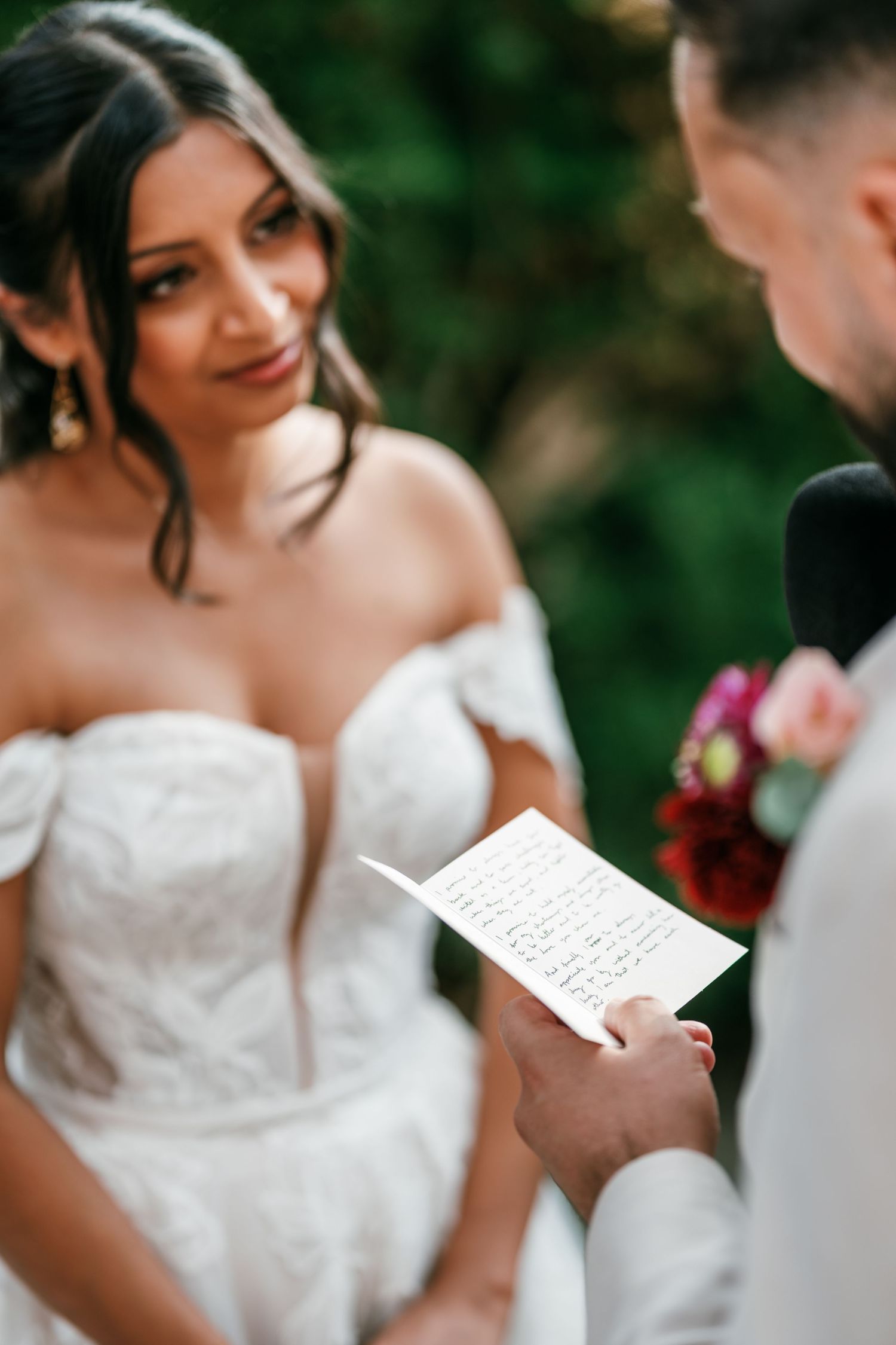Smiling bride in an off-the-shoulder lace wedding gown listens as the groom reads handwritten vows during an outdoor ceremony.