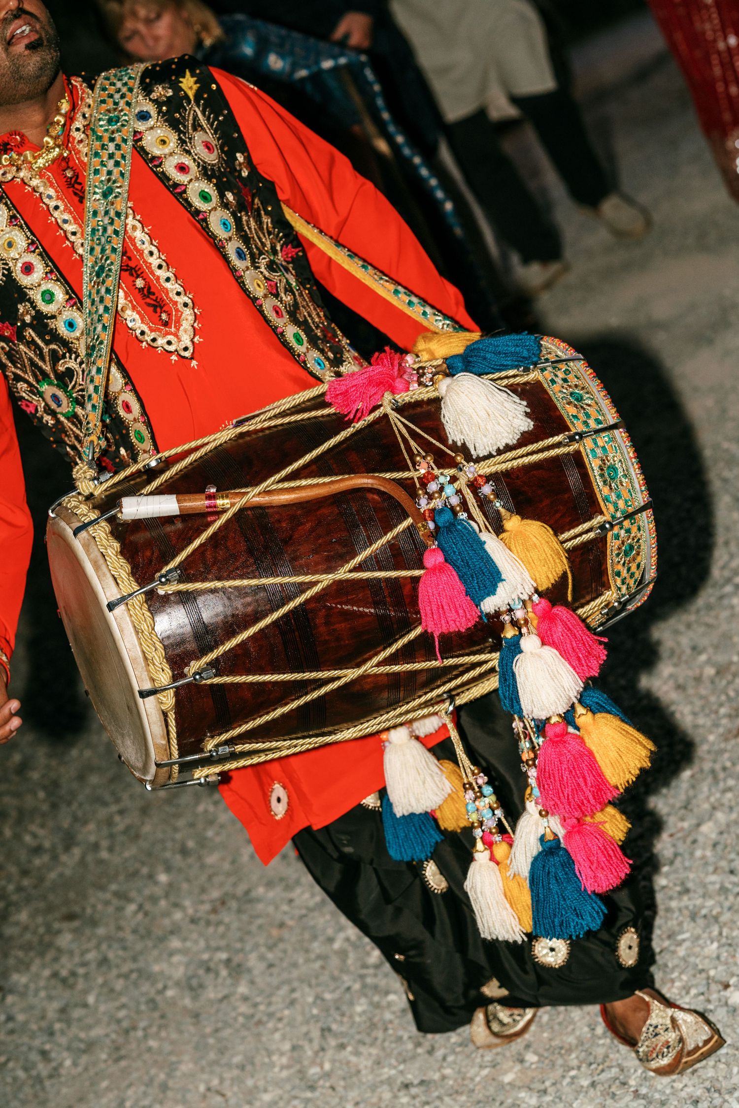 Musician in an orange embroidered tunic plays a wooden dhol drum decorated with colorful tassels during a festive South Asian celebration.