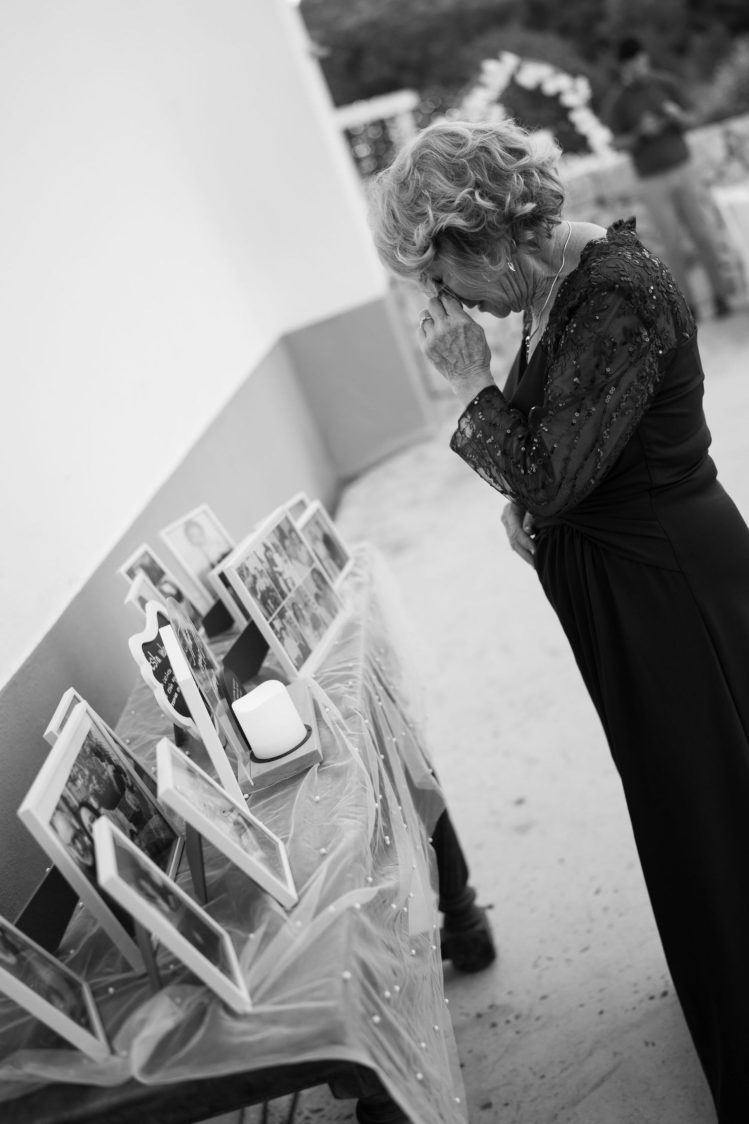 Black and white photo of an emotional woman in a lace dress wiping her eyes beside a memorial table of framed photos and a candle.