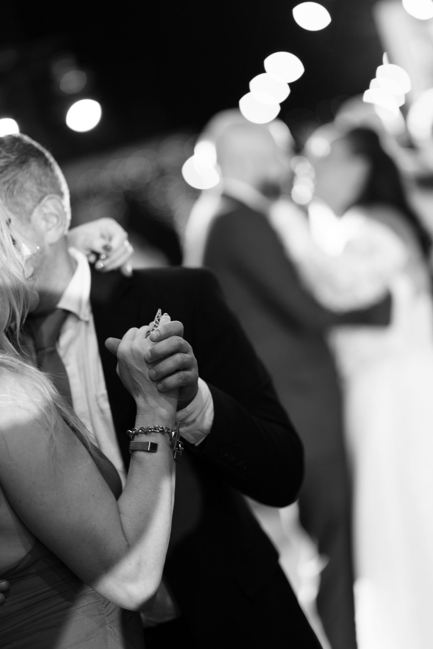 Black and white photo of two dancers holding hands close up at a wedding reception while another couple dances blurred in the background.