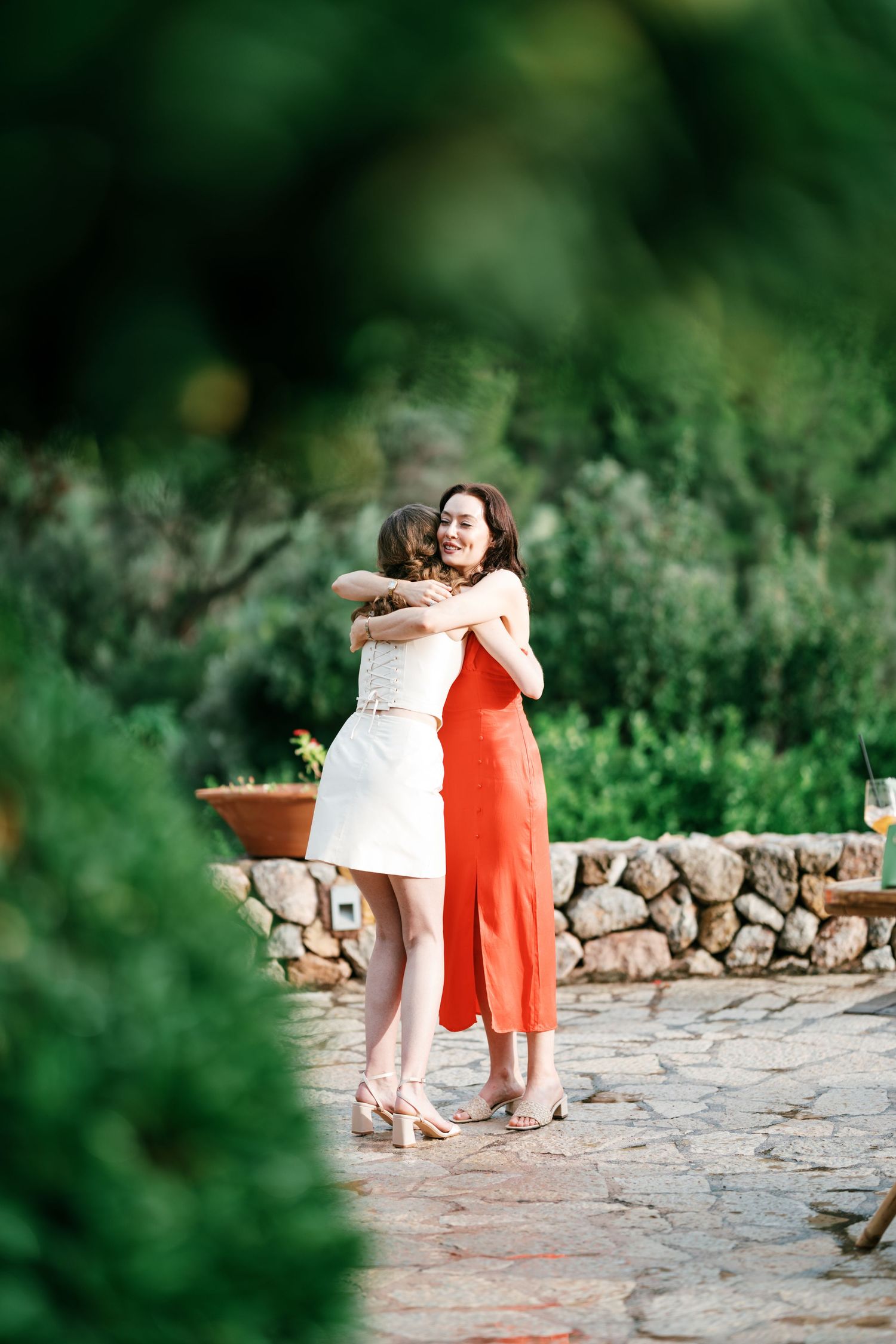 Two women embrace in a warm hug on a stone patio, one in a white dress and one in an orange dress, surrounded by greenery.