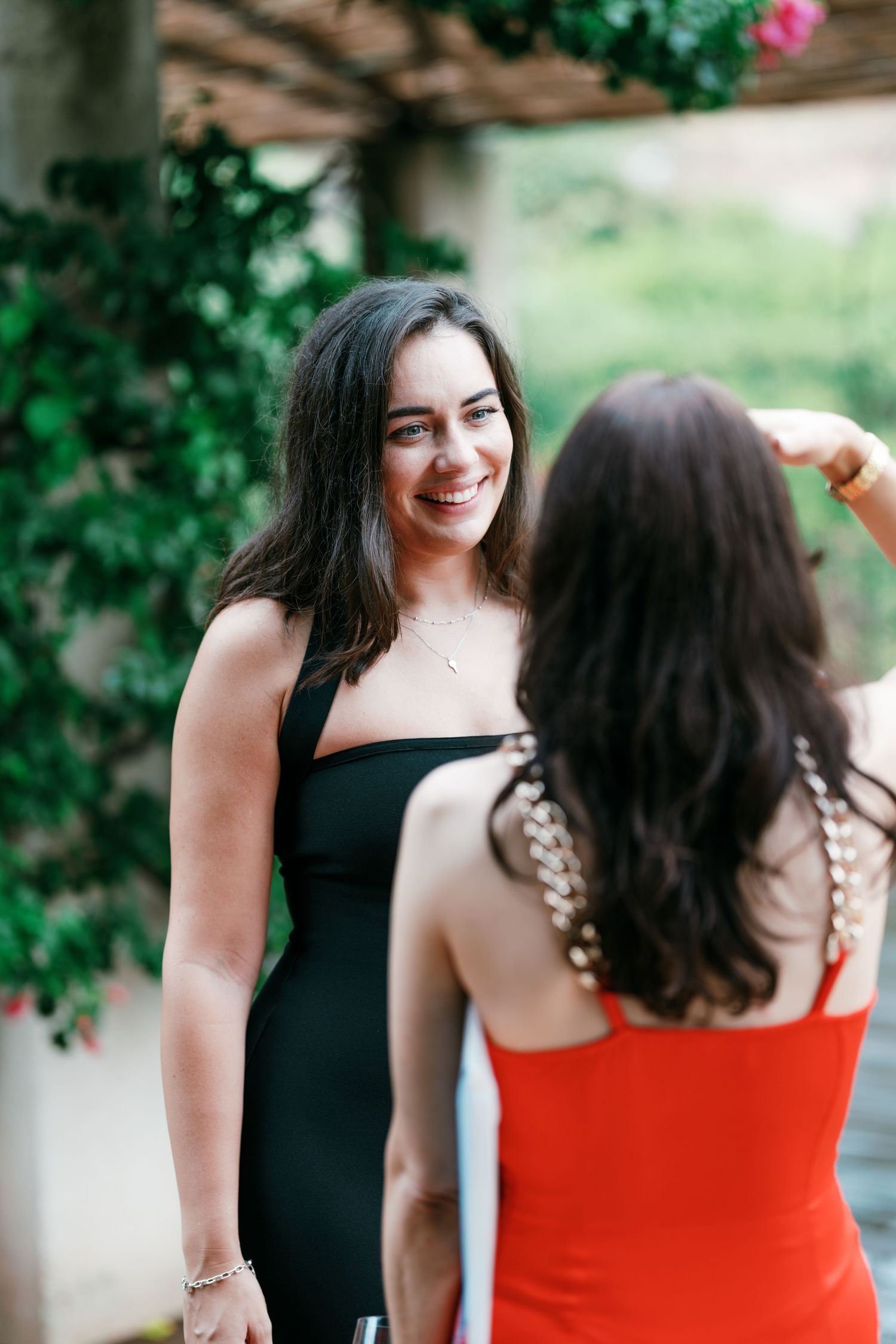 Smiling woman in a black dress chats with a woman in a red dress on a vine-covered terrace at an outdoor event.