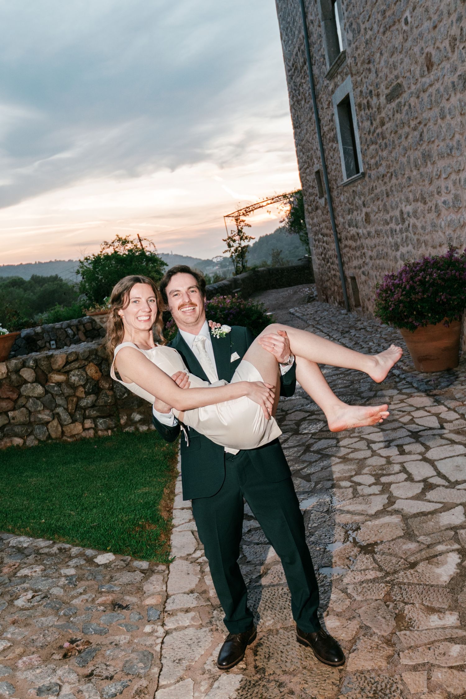 Groom in a dark green suit carries a barefoot bride in a white dress along a stone path at sunset beside a rustic stone building.