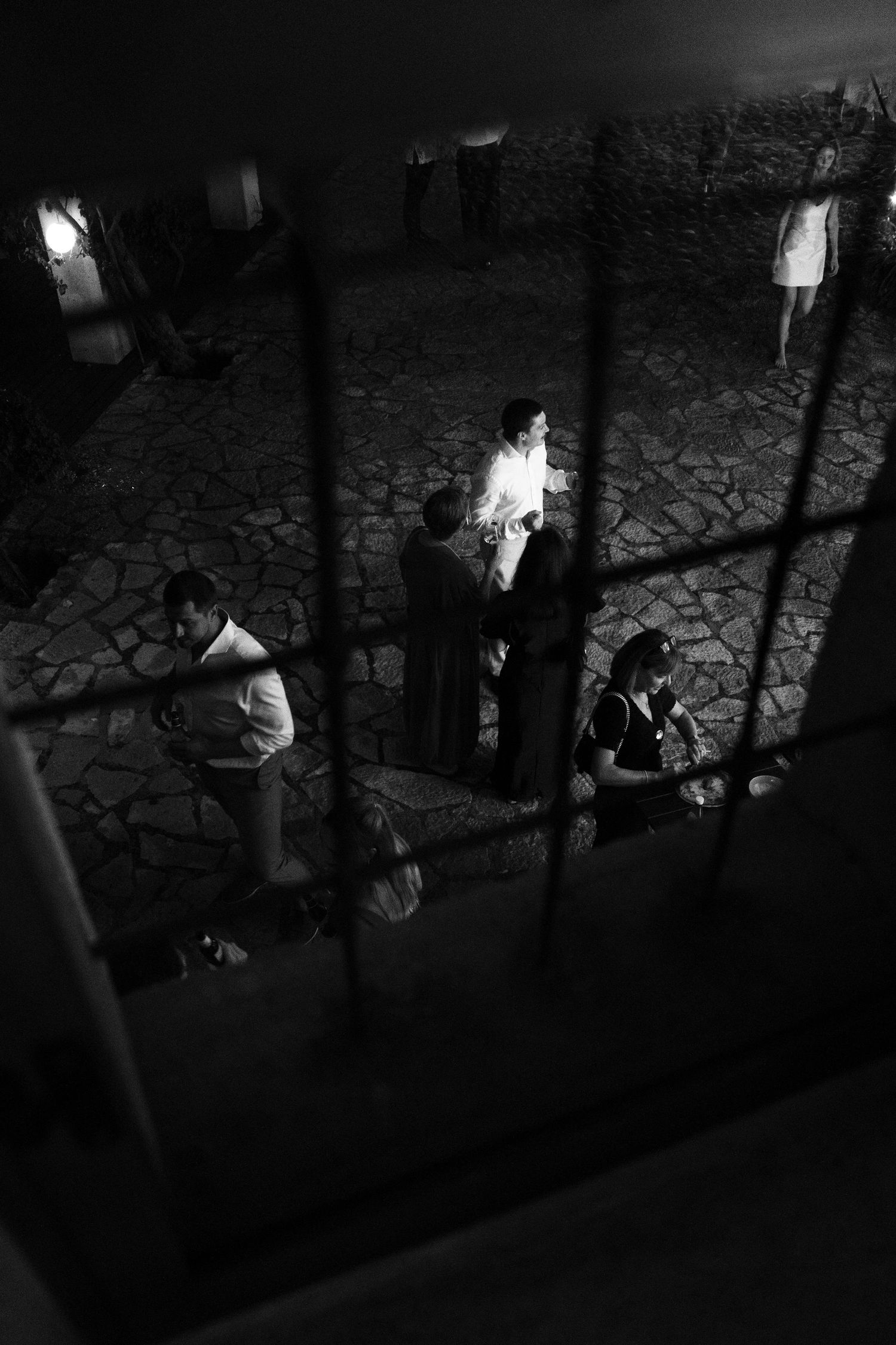 Black and white view through a window grid of wedding guests mingling on a dark cobblestone courtyard at night.