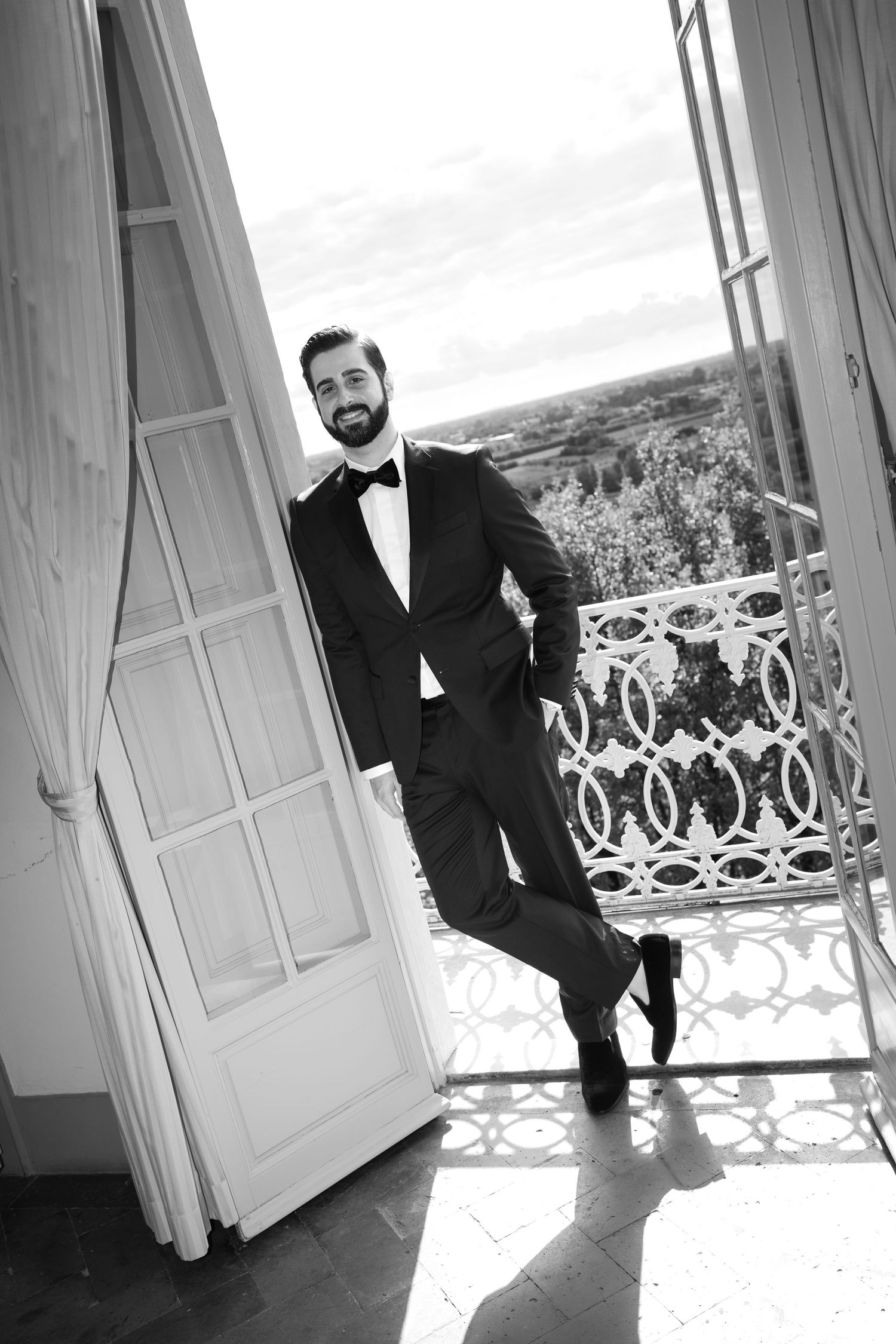 Bearded man in a tuxedo and bow tie leans against an open balcony door with an ornate railing and scenic landscape view, in black and white.