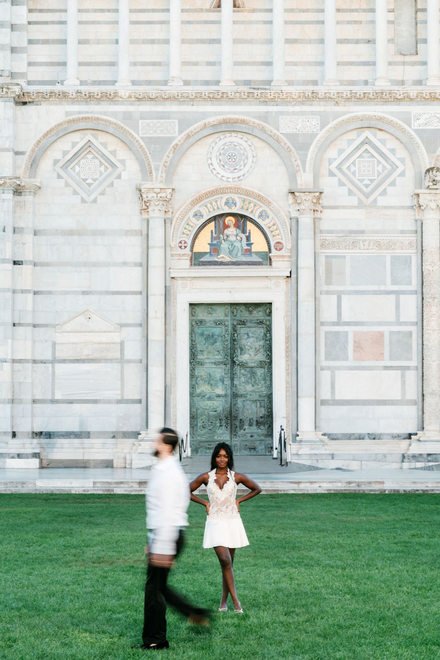 Woman in a white dress poses on green grass before a marble cathedral with a green door while a blurred man walks past.