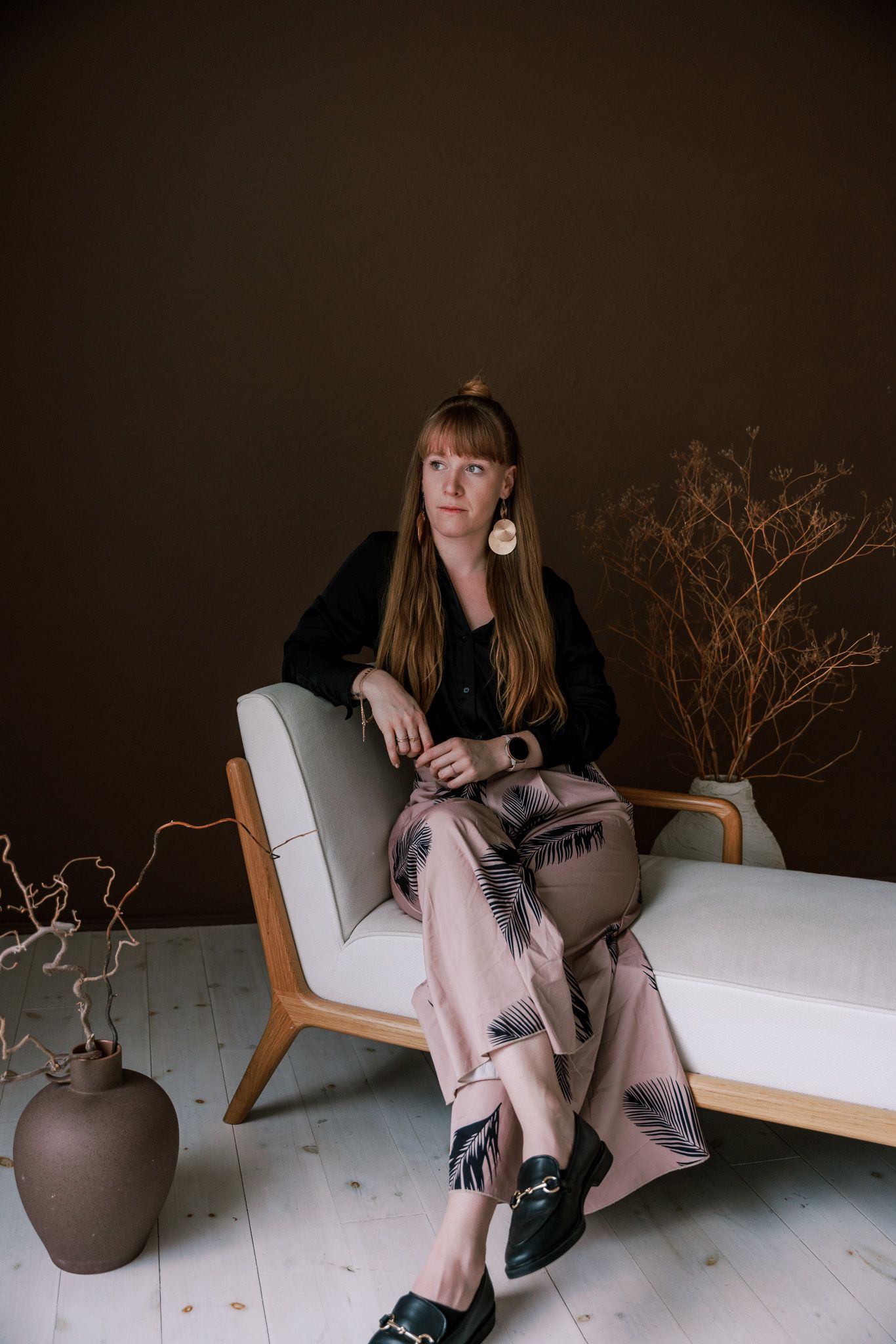 Elegant woman in black blouse and palm-print trousers seated on a modern chaise lounge in a dark studio. Mallorca wedding photographer, film