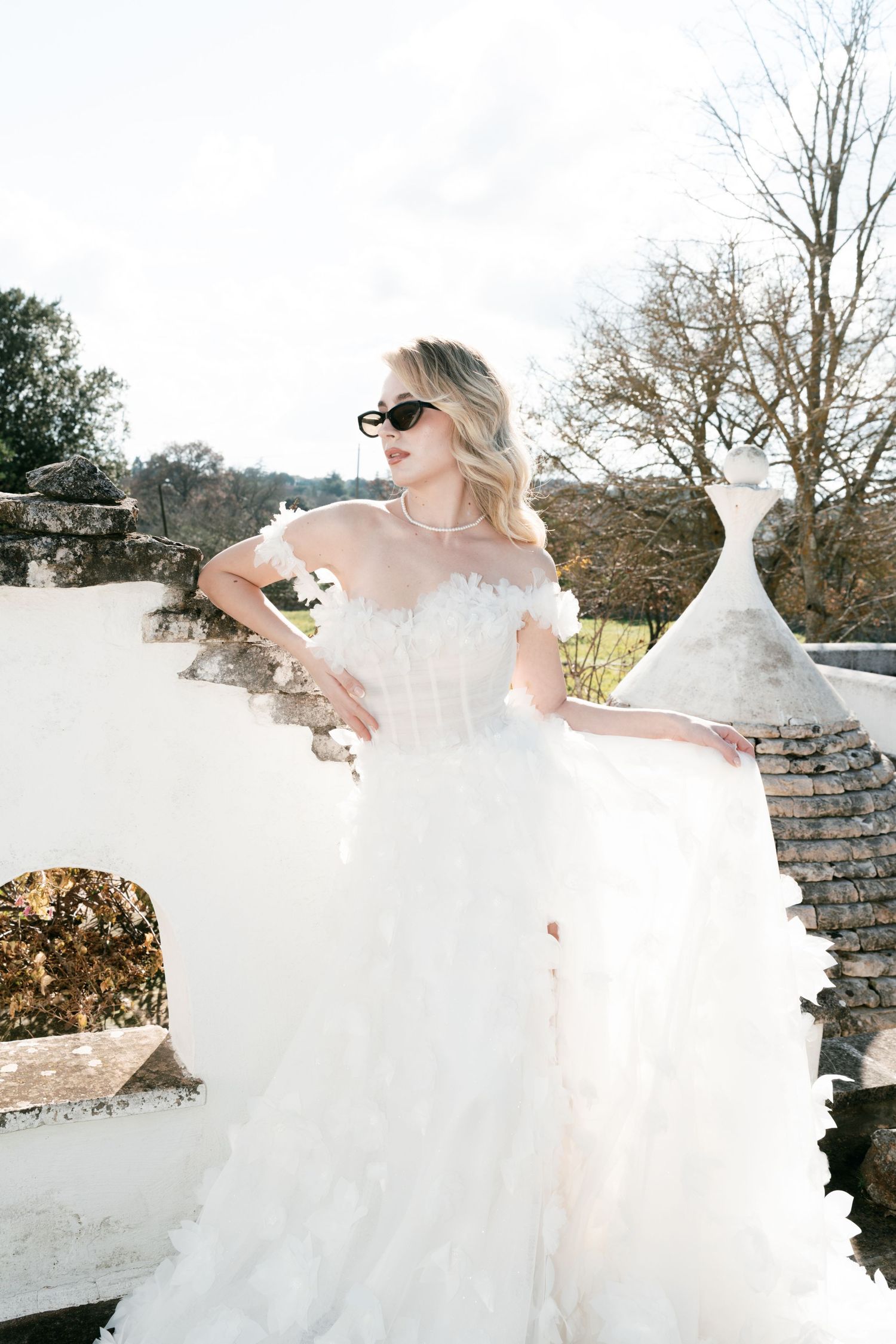 Blonde bride in a white ruffled strapless gown wearing sunglasses on a sunlit stone terrace. Puglia wedding photographer, film