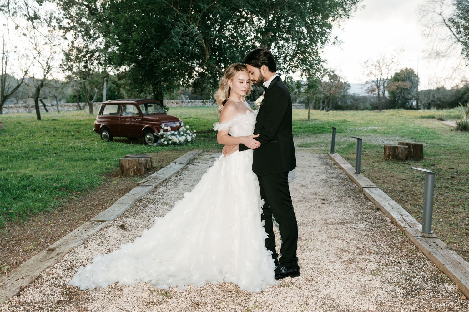 Bride and groom embracing on a gravel path with a vintage red car and olive trees behind them. Puglia wedding photographer, film