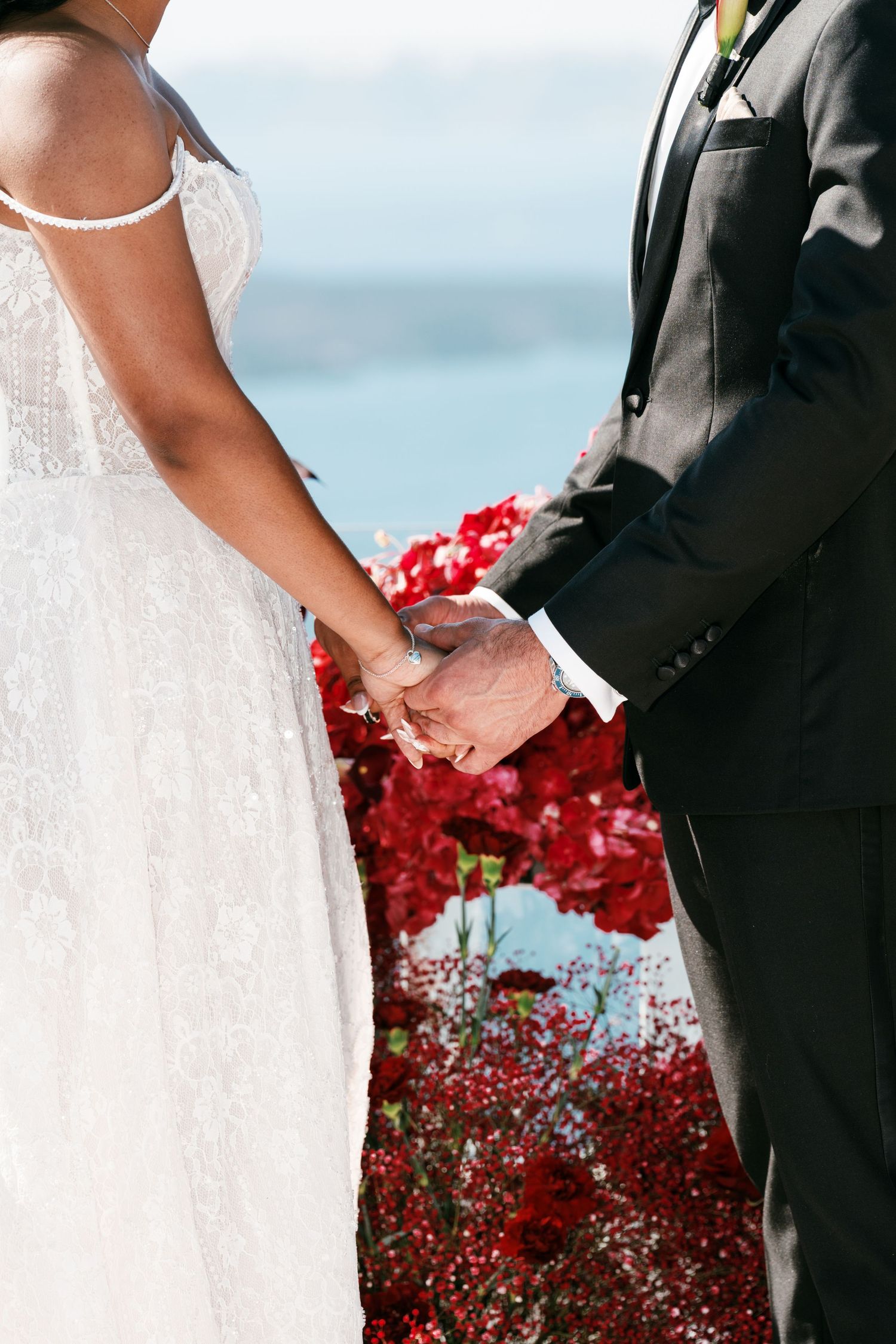 Close-up of bride and groom holding hands at wedding ceremony with vibrant red flowers in background.