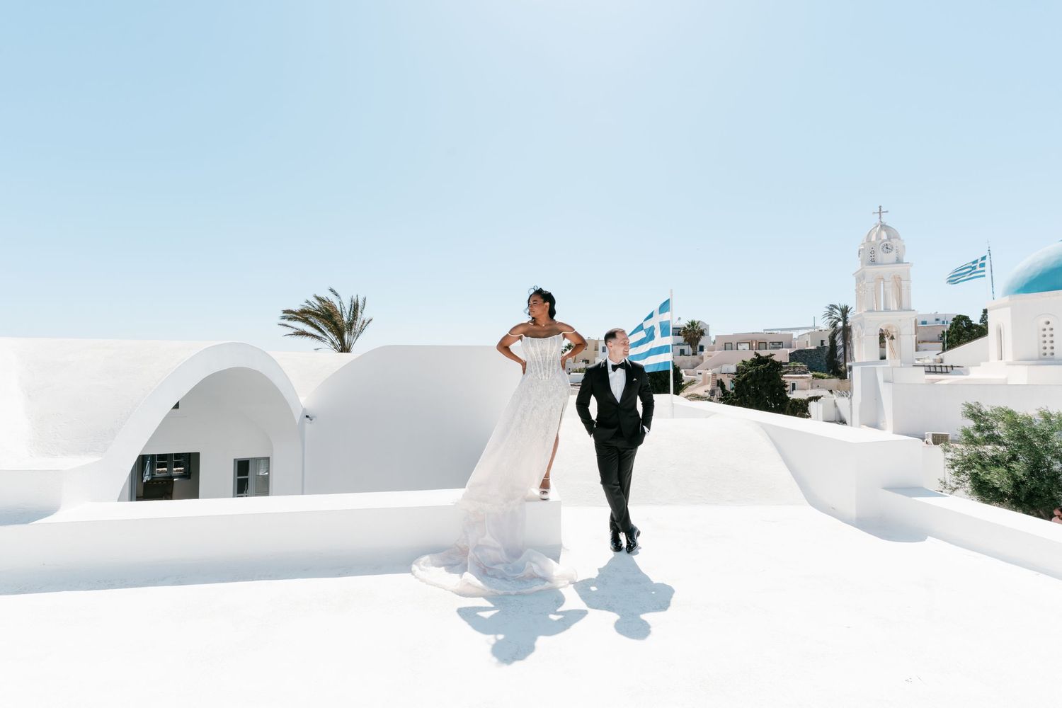 Bride and groom walking on white rooftop in Santorini Greece with blue-domed church and Greek flag in background.