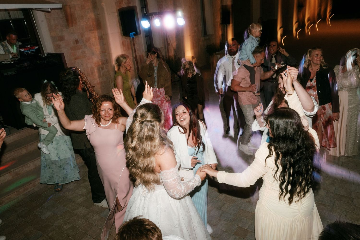 Guests dancing joyfully at a wedding reception, with a bride in white gown surrounded by friends under colorful lights in Mallorca.