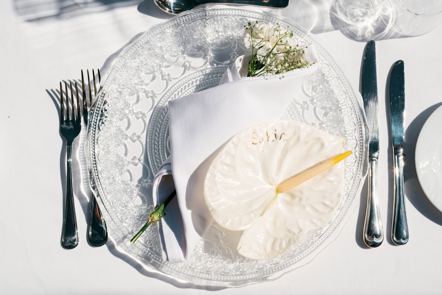 Elegant wedding table setting with silver charger plate, white napkin, floral decor, and polished silverware on white tablecloth.