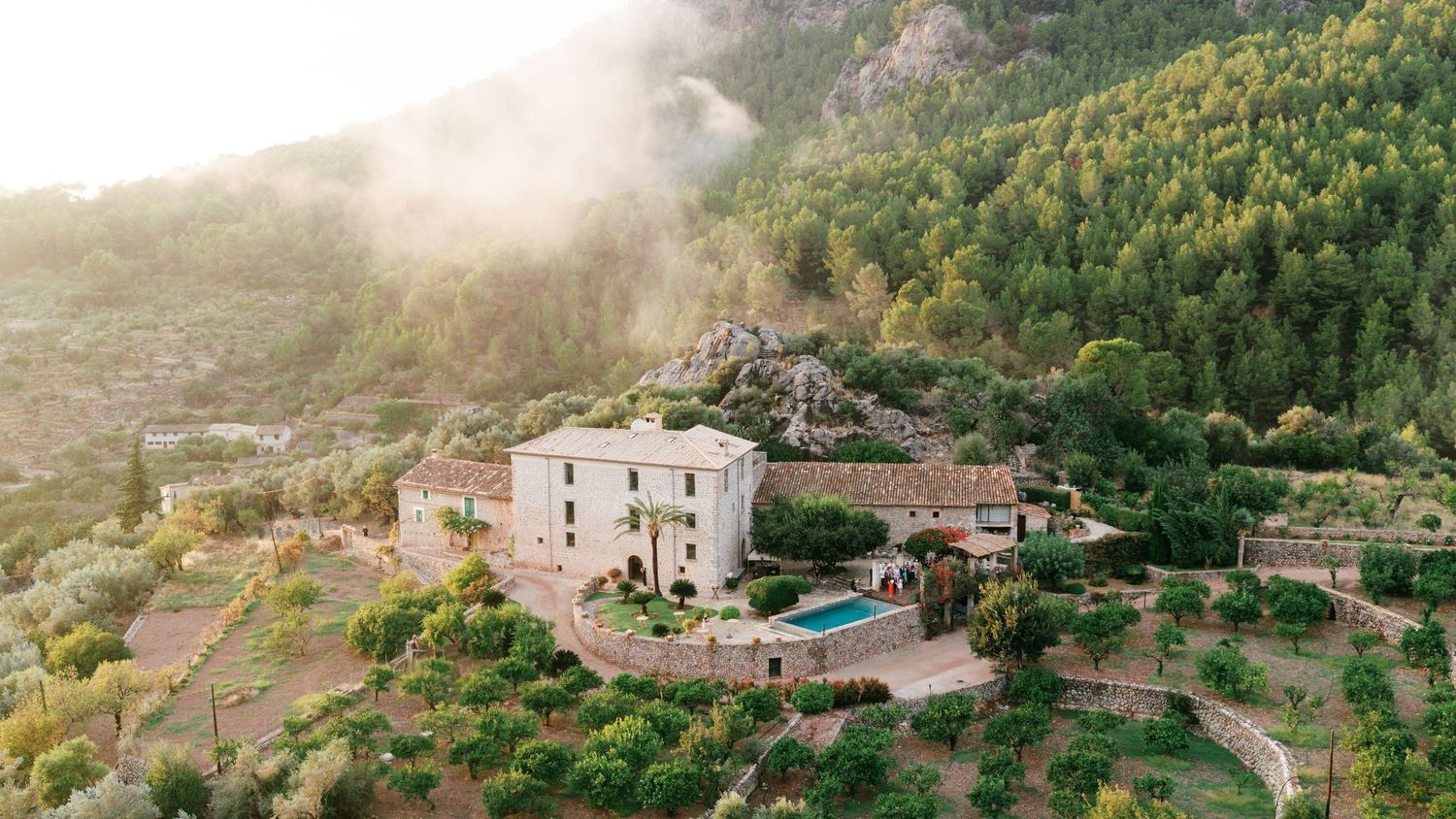 Aerial view of a stone villa with a swimming pool surrounded by terraced olive groves and misty forested mountains at sunrise.