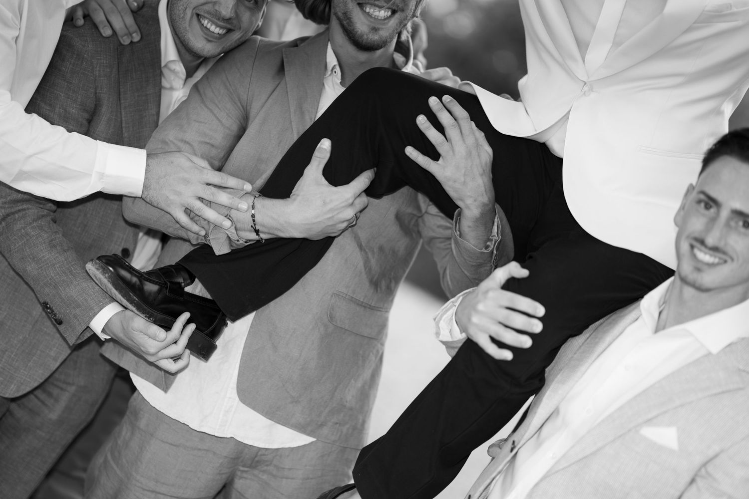 Groomsmen in suits joyfully lifting the groom in a black-and-white wedding celebration photo.