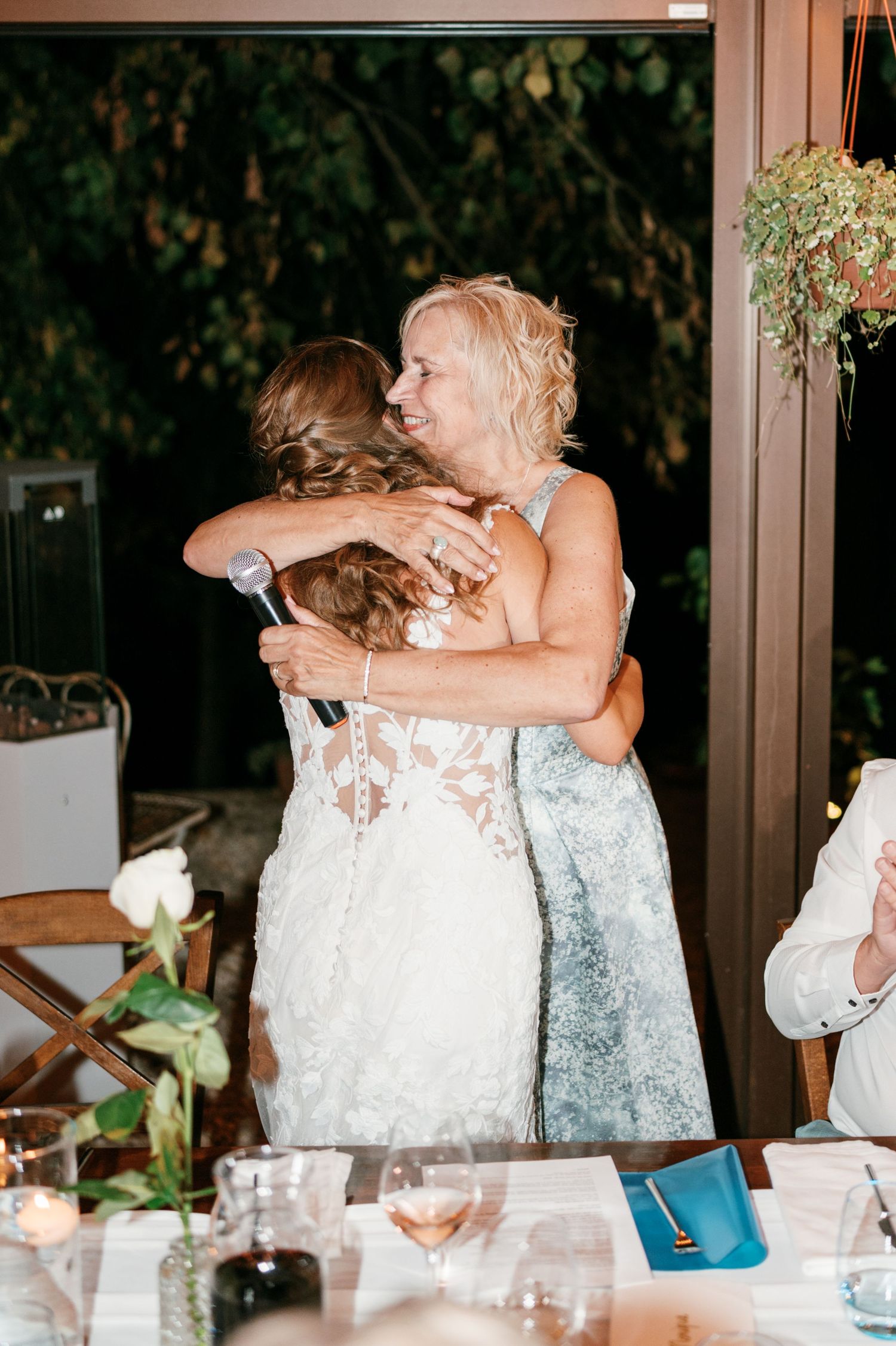 Bride in lace gown shares warm hug with older woman holding microphone at wedding.