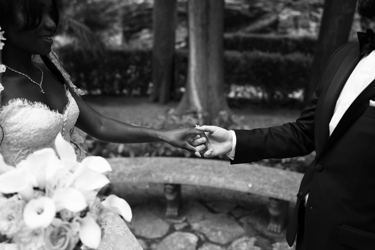 Black and white photo of a bride in a lace gown holding hands with a groom in a tuxedo, with a white floral bouquet in a garden.