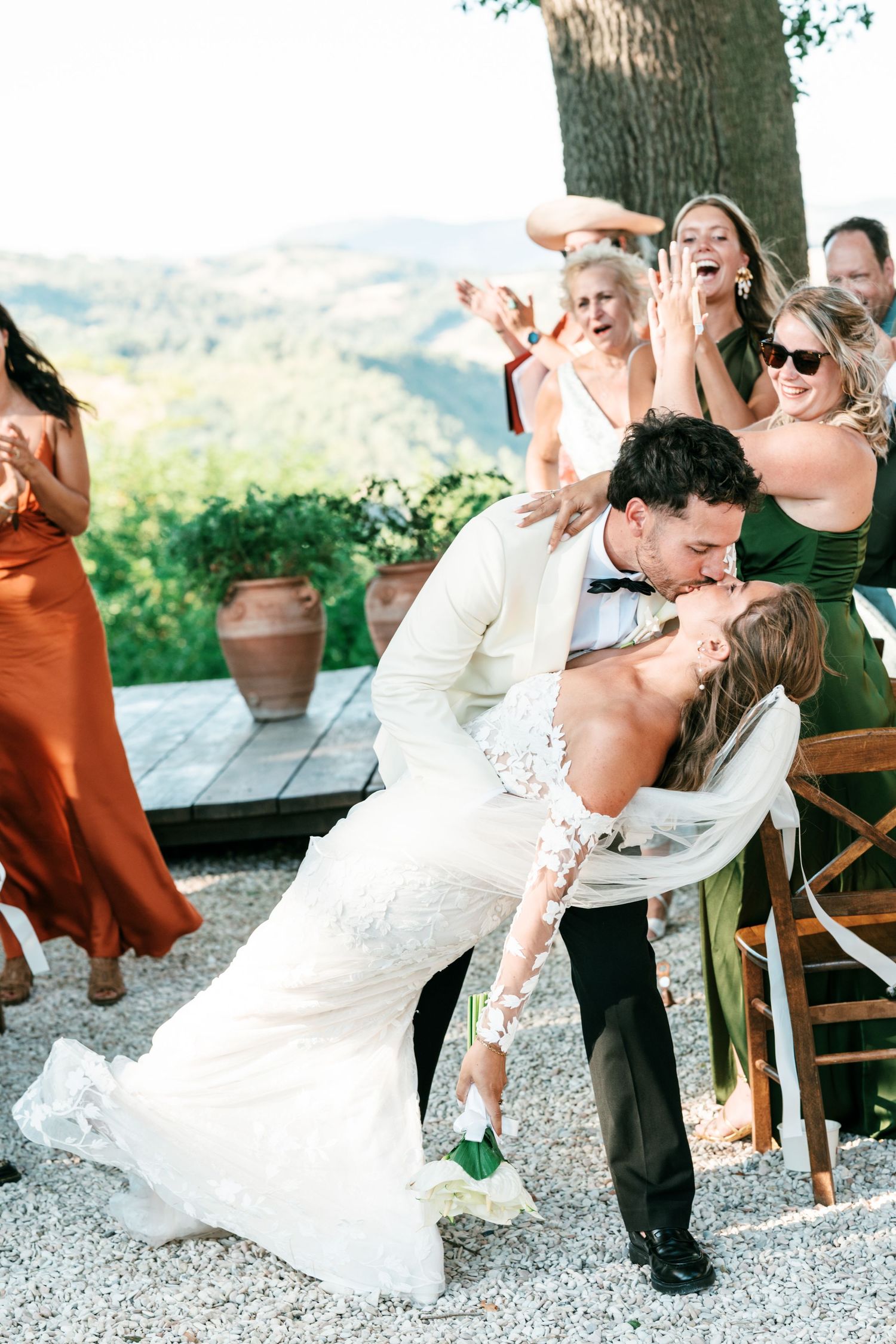Groom dips bride for a romantic kiss at outdoor Tuscan wedding while bridesmaids in green and rust dresses cheer.