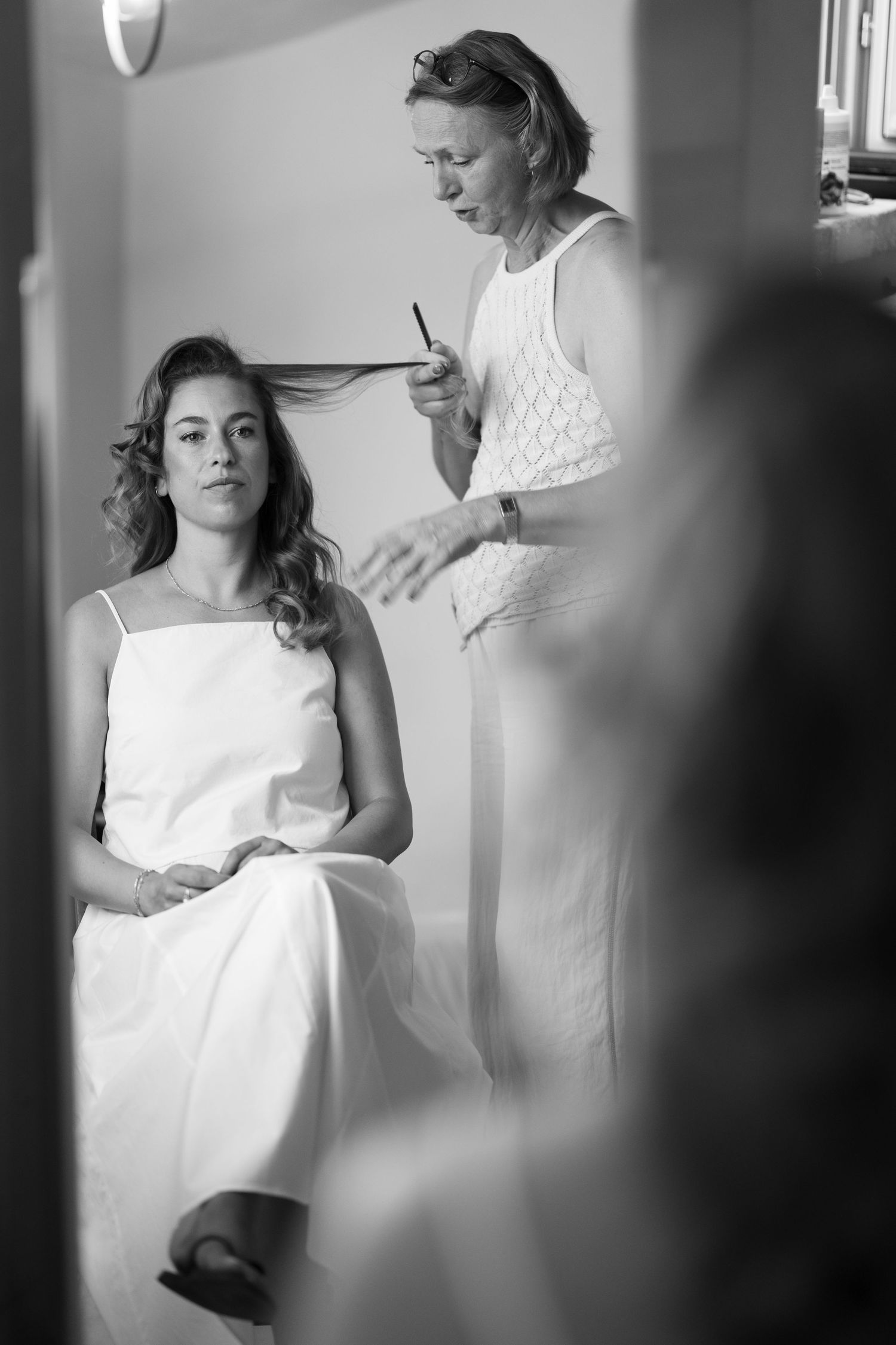 Black and white photo of a bride in white dress getting hair styled by a hairdresser before her wedding.