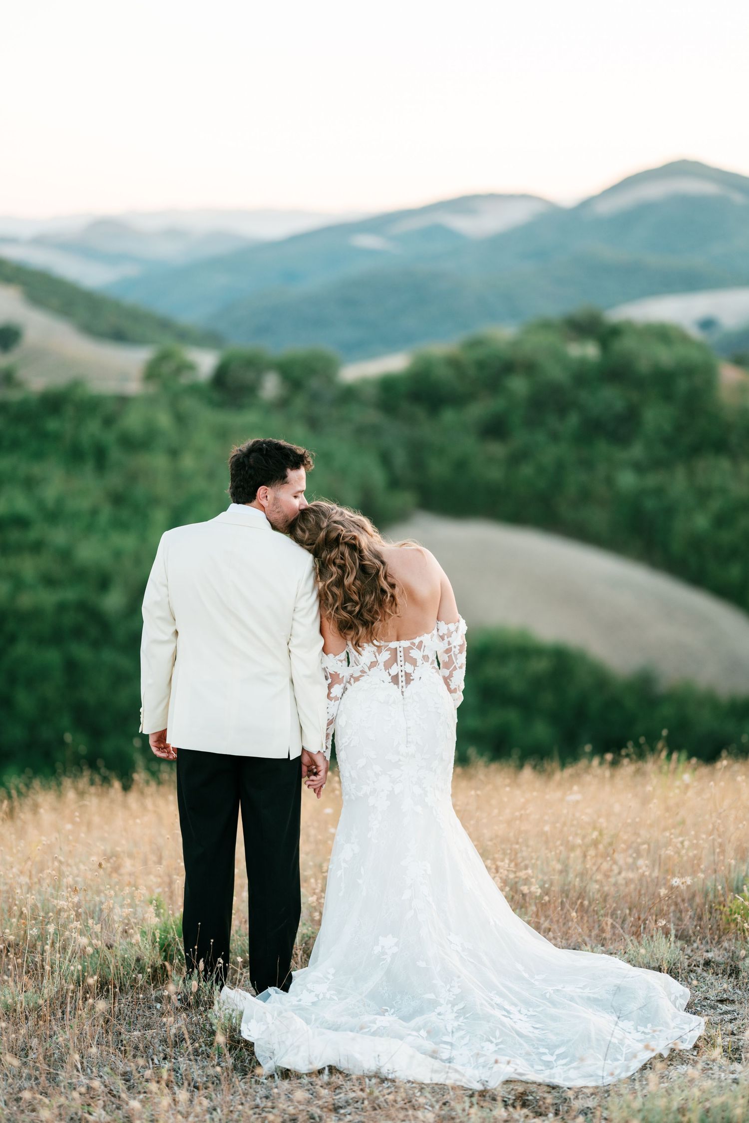 Bride and groom holding hands overlooking rolling green hills at golden hour, bride in lace off-shoulder gown with train.