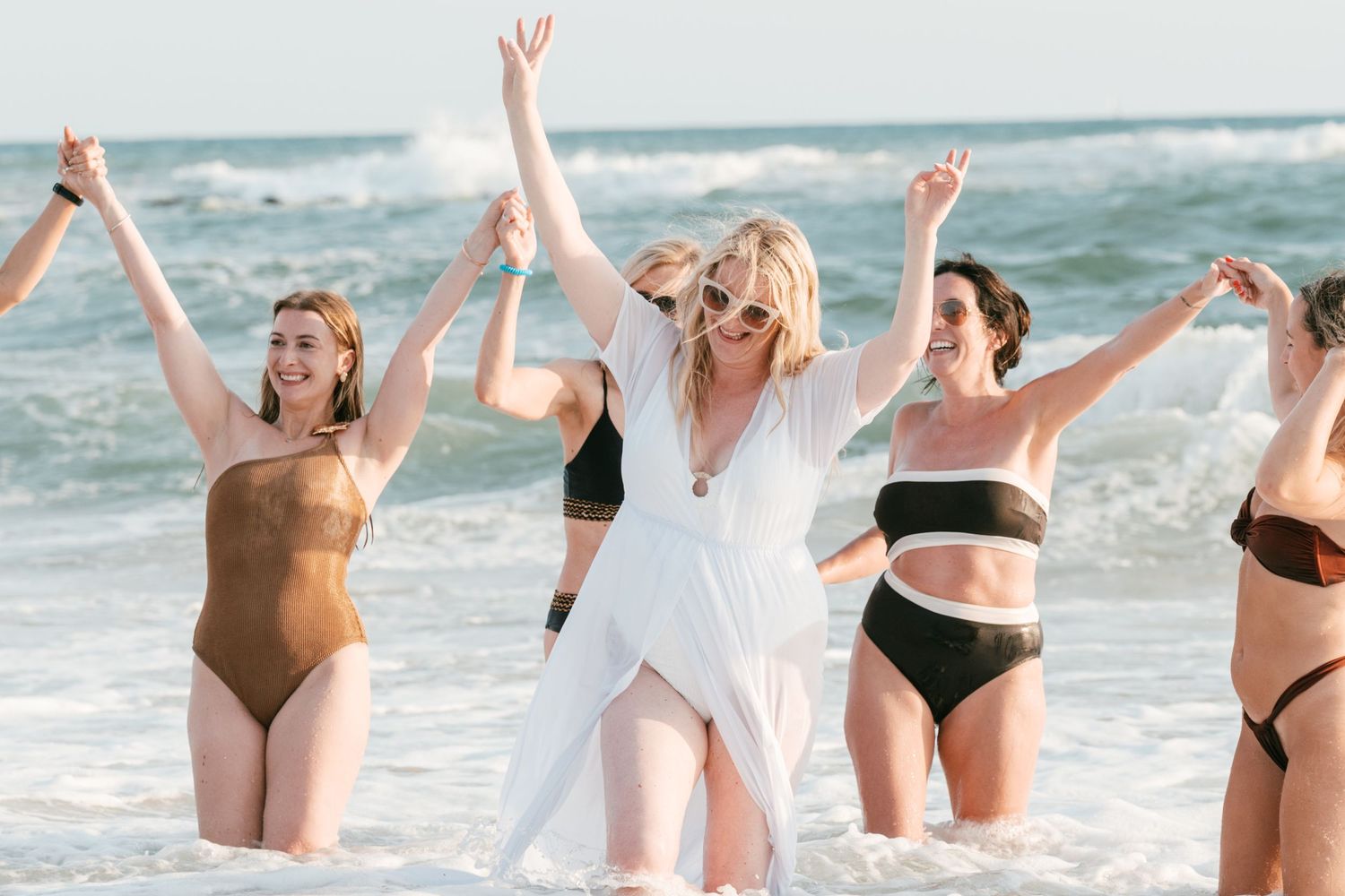 Group of women in swimsuits celebrating on a beach, splashing in ocean waves with arms raised joyfully. Mallorca wedding filmmaker, video