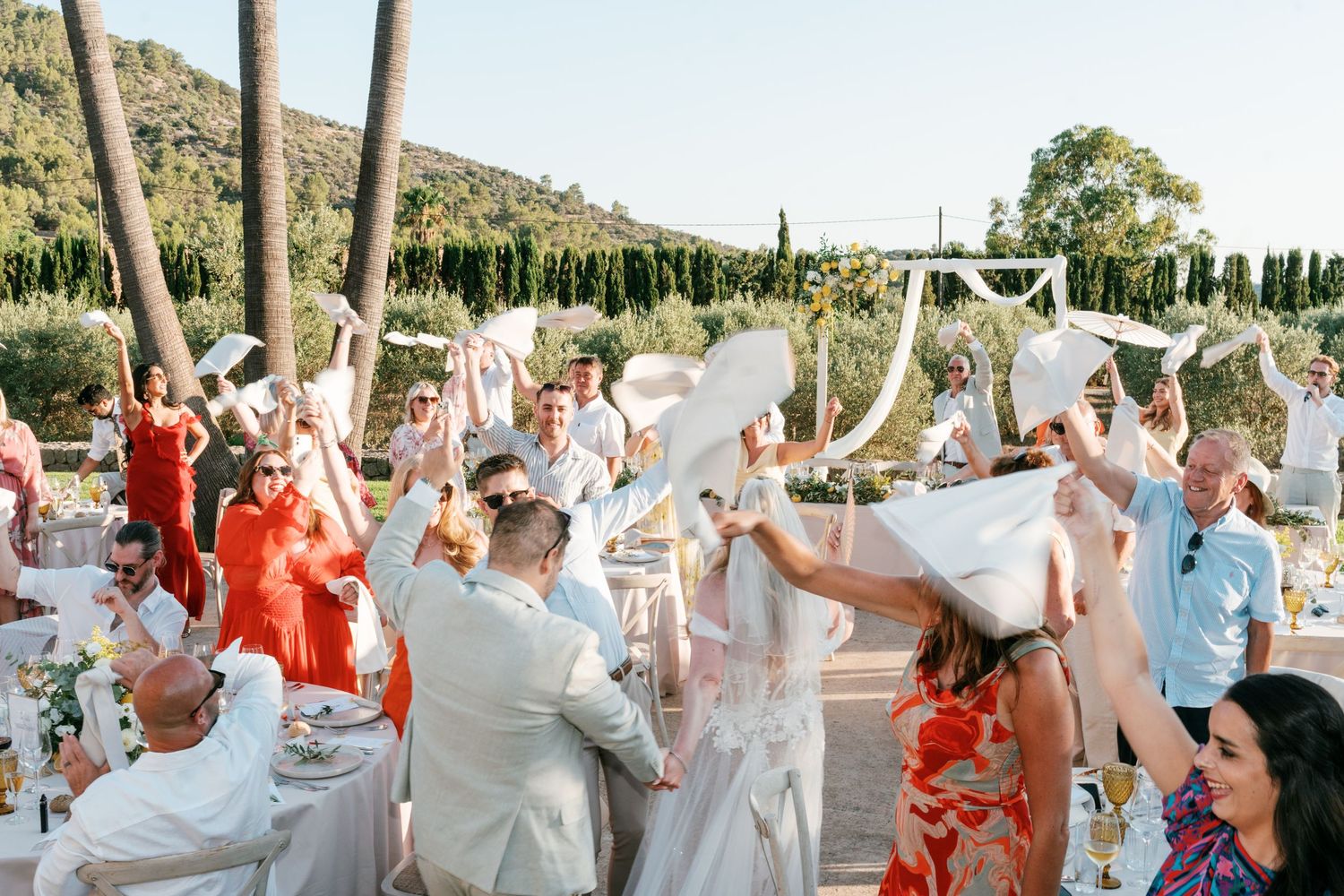 Wedding guests celebrate outdoors waving white napkins at a sunny reception in a scenic garden. Mallorca wedding photographer, filmmaker