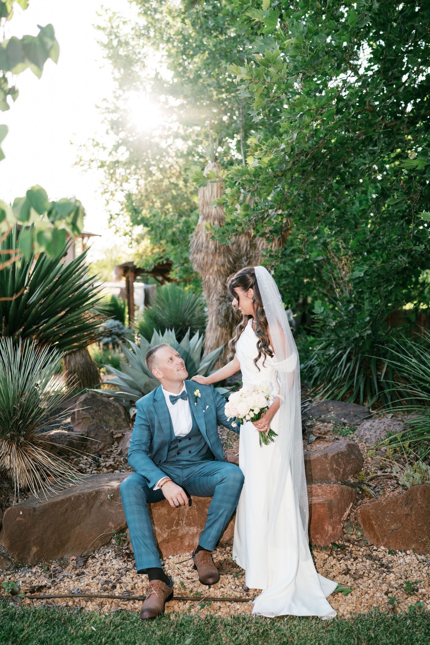 Bride in white gown and veil holds bouquet while groom in blue suit sits on stone wall in Castello di Vicarello, Tuscany.
