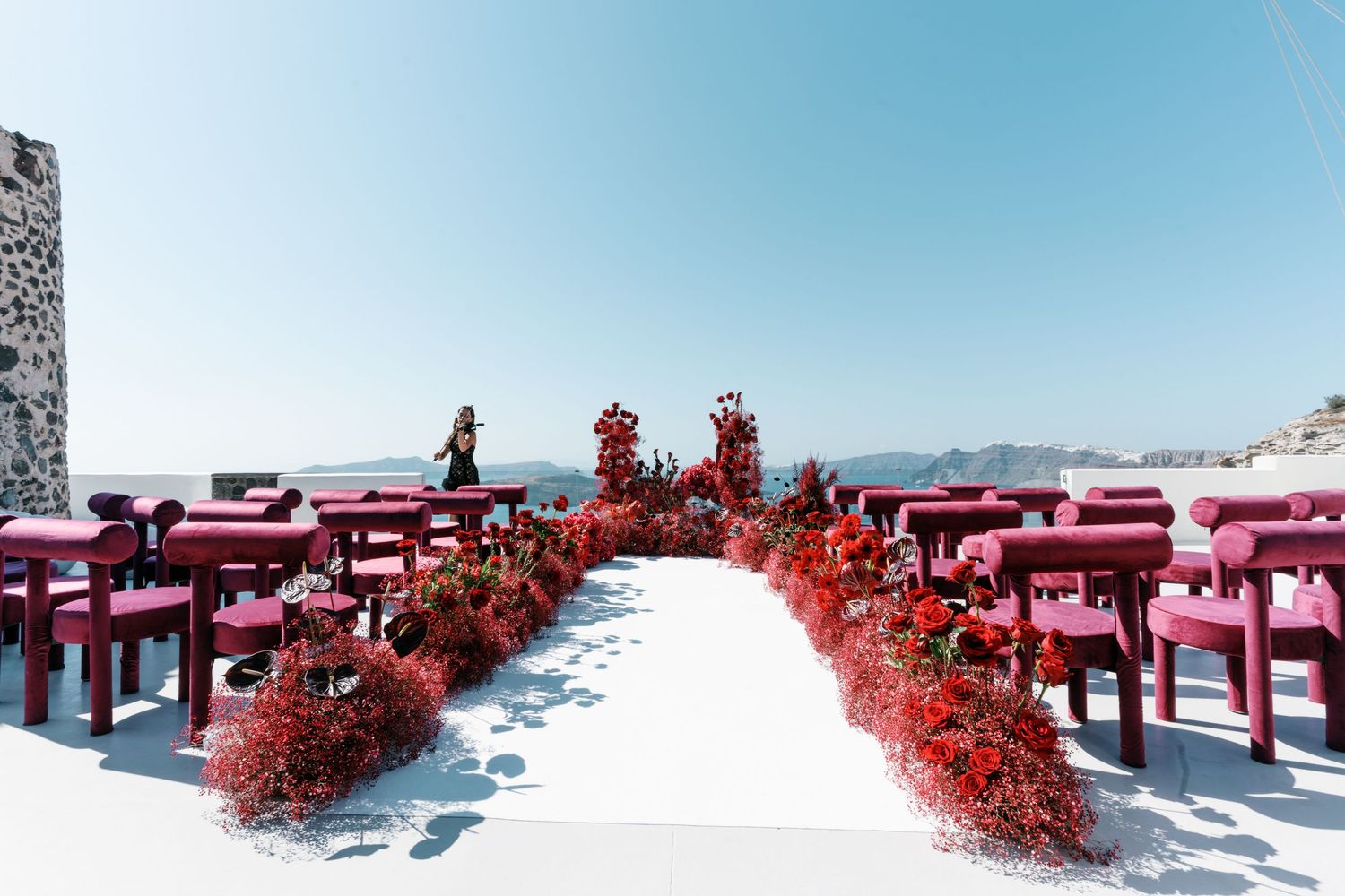 Outdoor wedding ceremony setup in Santorini with red floral aisle, pink chairs, and stunning caldera views under clear blue sky.