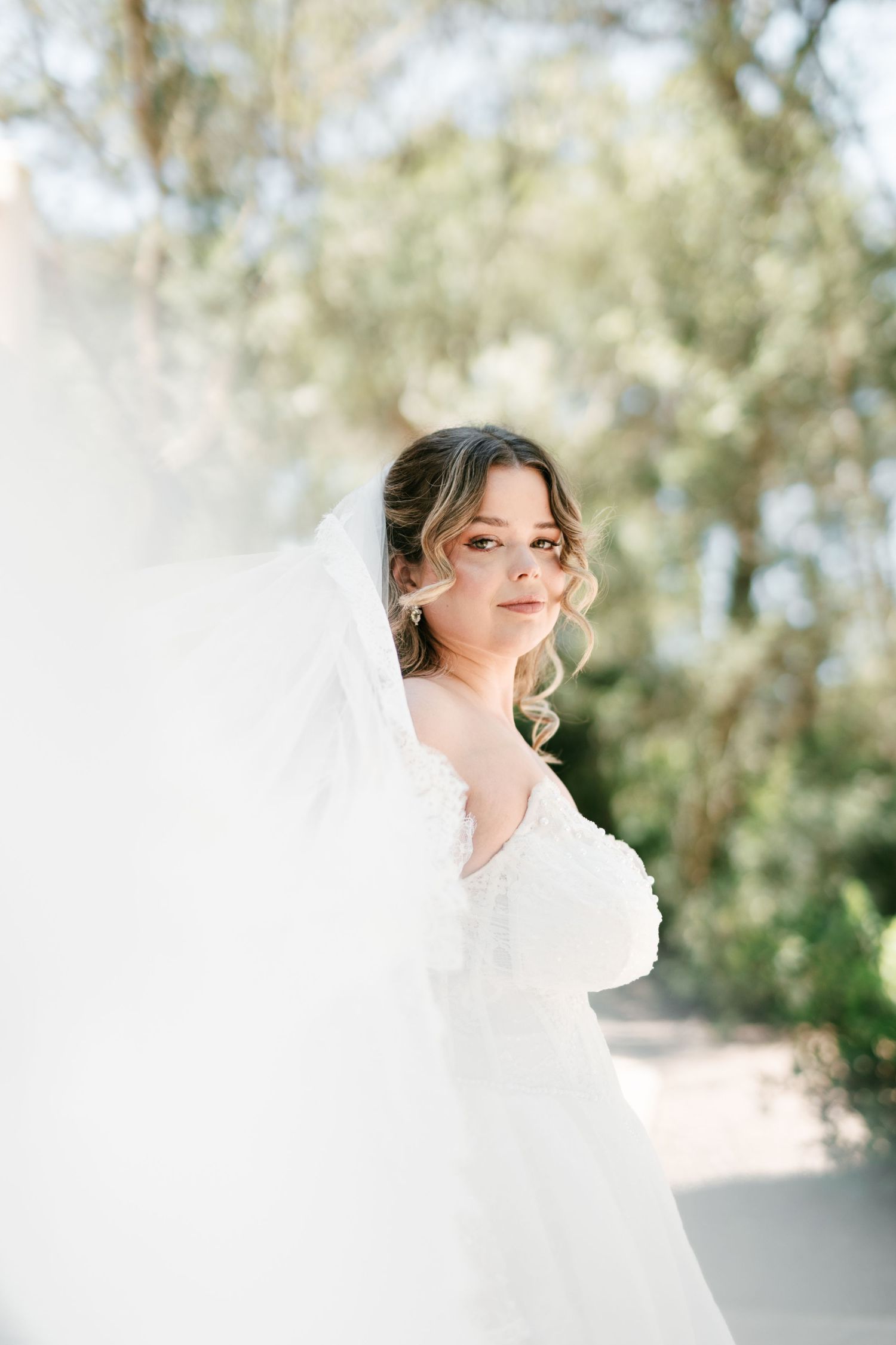 Bride in white wedding gown and flowing veil stands outdoors among lush green trees in soft natural light.