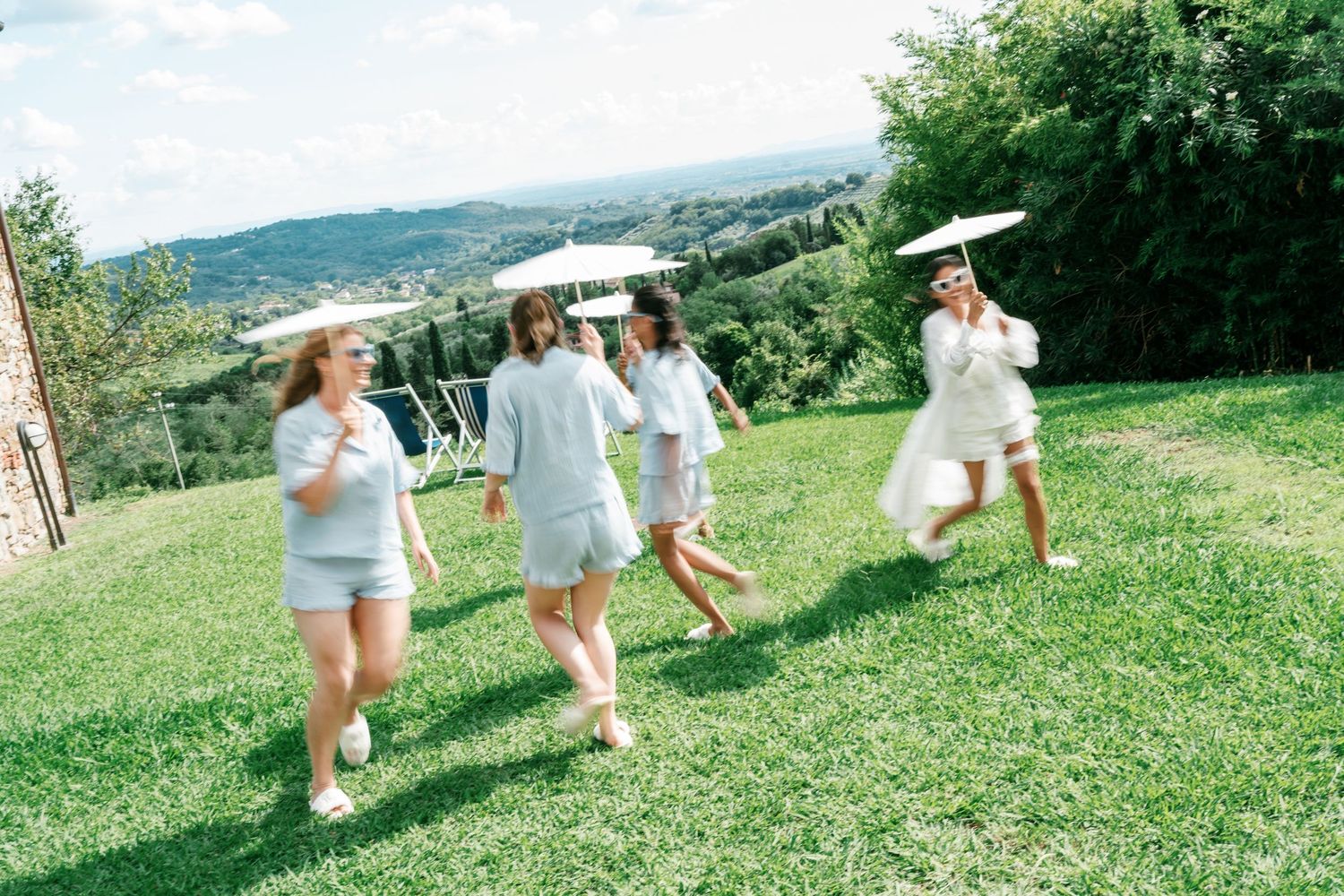 Bridesmaids in matching light robes and shorts dance with white parasols on a sunny grassy hillside overlooking green Tuscan countryside.