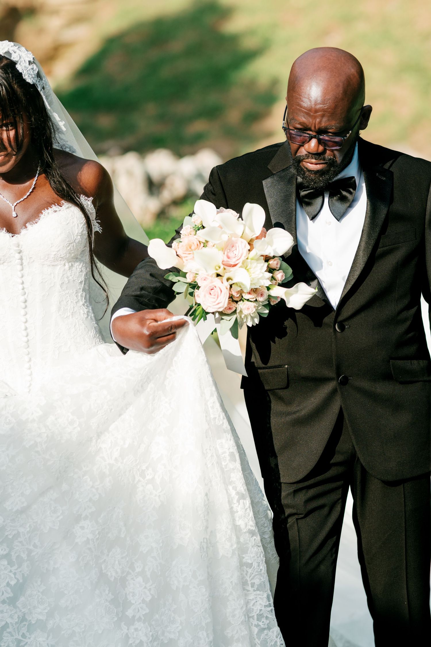 Father in a black tuxedo and bow tie walks his daughter, a bride in a lace wedding gown and veil, holding a pink and white bouquet outdoors.