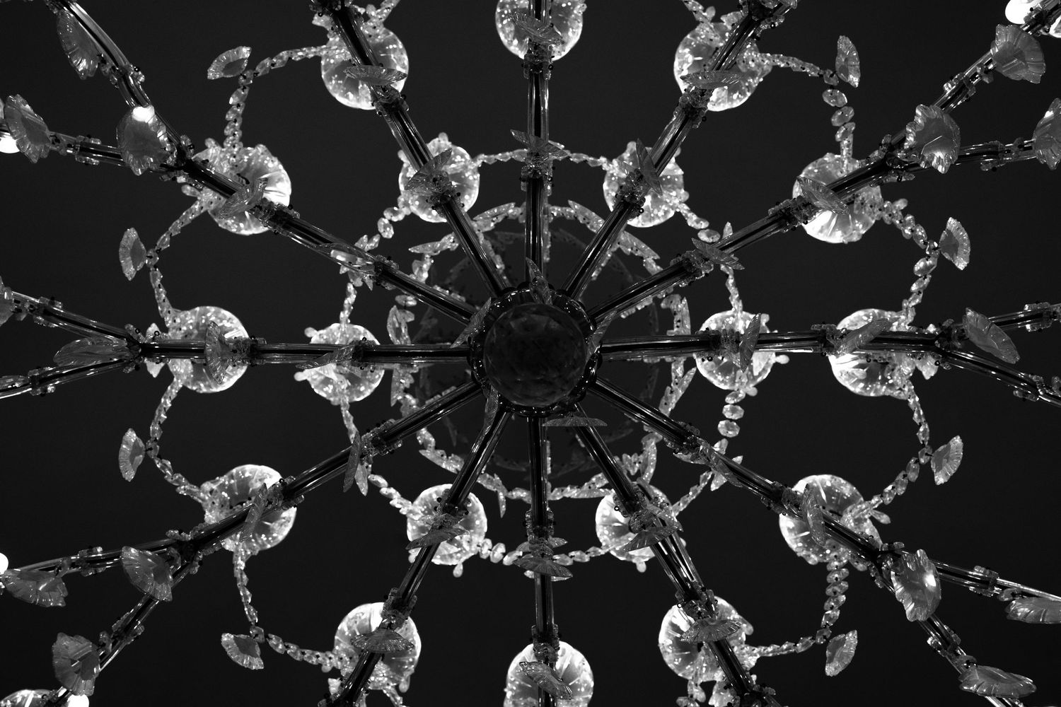 Black and white view looking up at an ornate crystal chandelier with radiating arms and glass droplets against a dark ceiling.