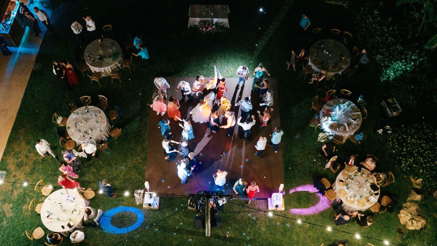 Aerial view of guests dancing on a lit dance floor surrounded by round banquet tables at an outdoor evening wedding reception on a lawn.