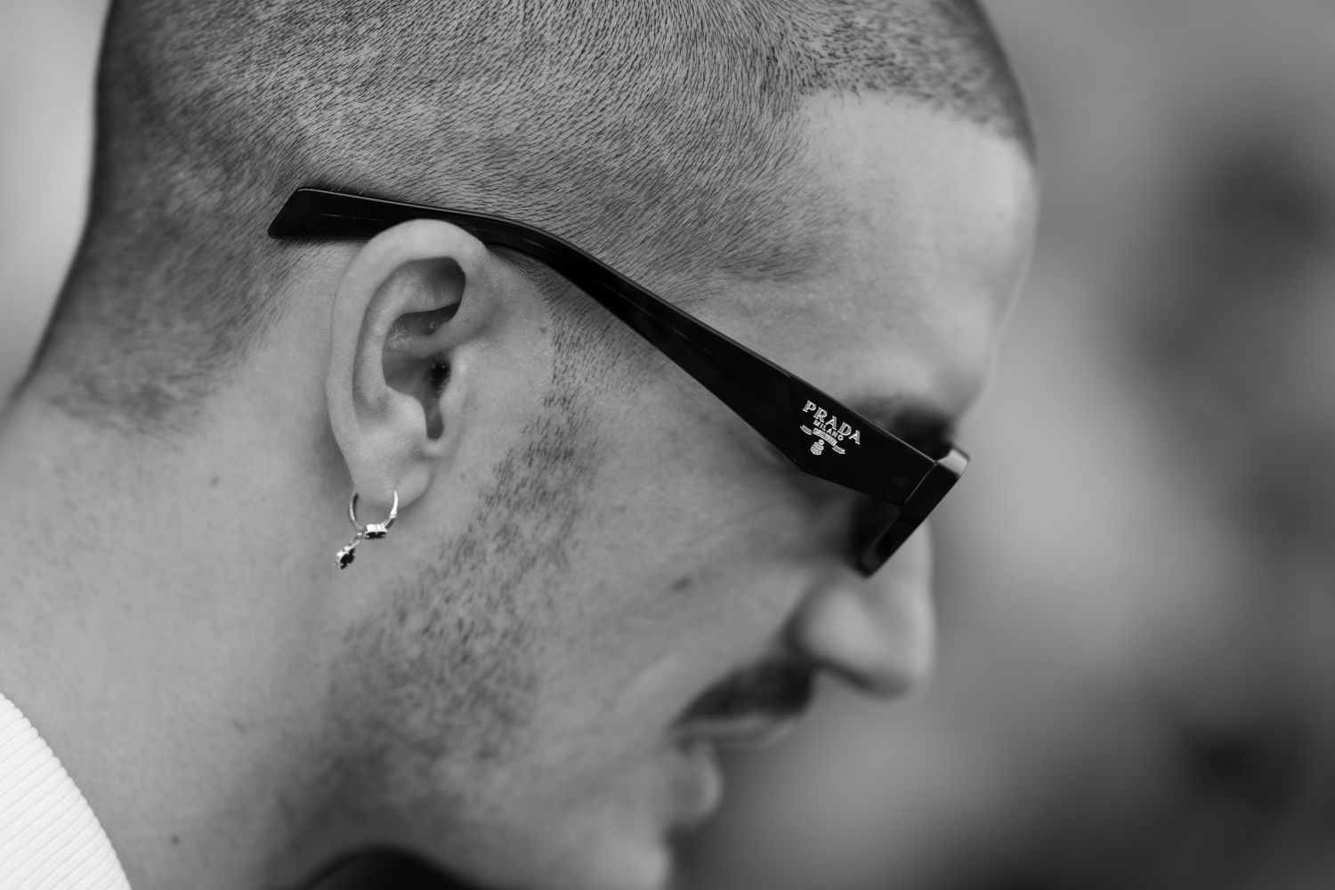 Black and white side profile of a man with short hair, stubble, Prada sunglasses and a small dangling earring.