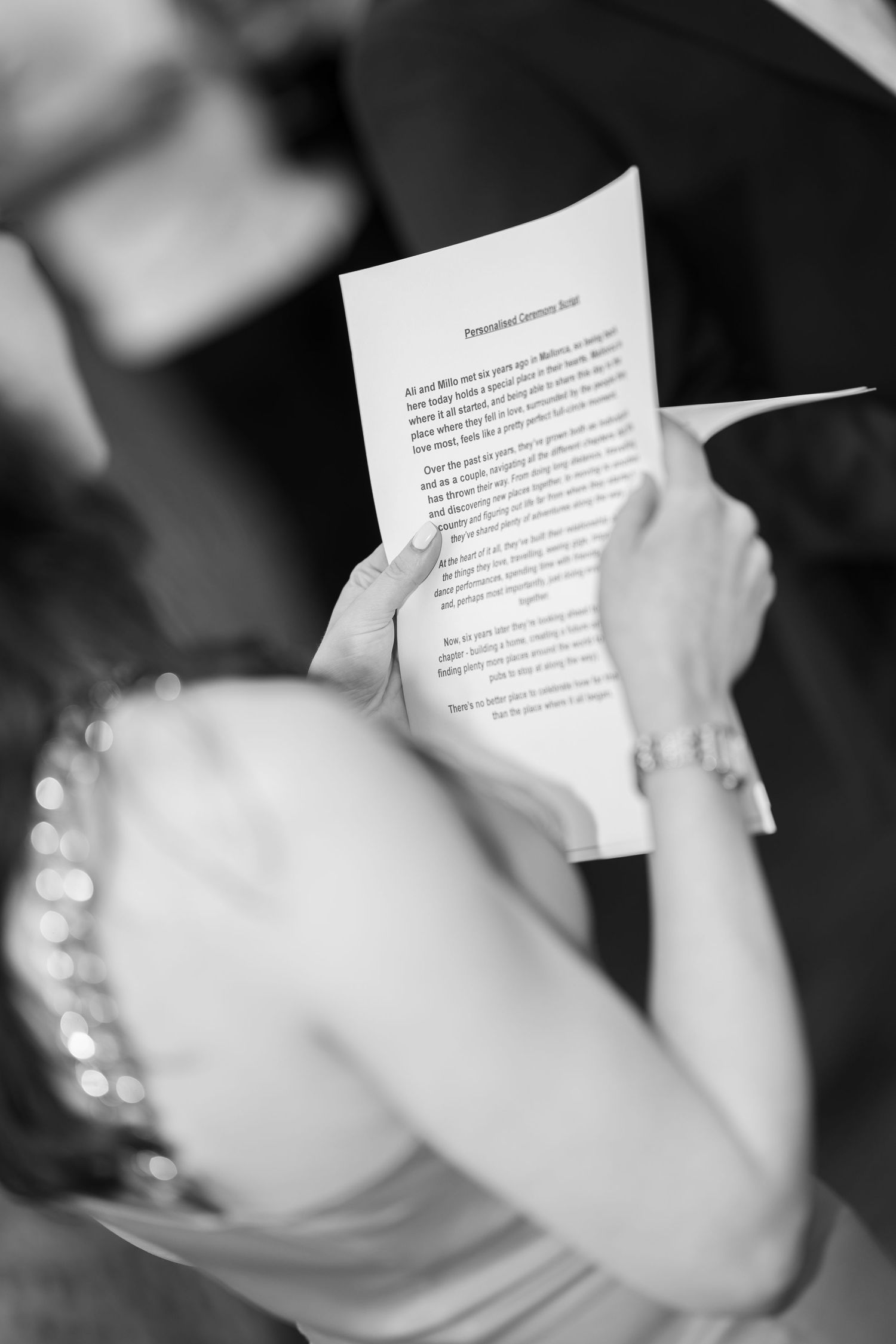 Woman in a beaded dress holds a printed wedding ceremony script in black and white during an outdoor celebration.