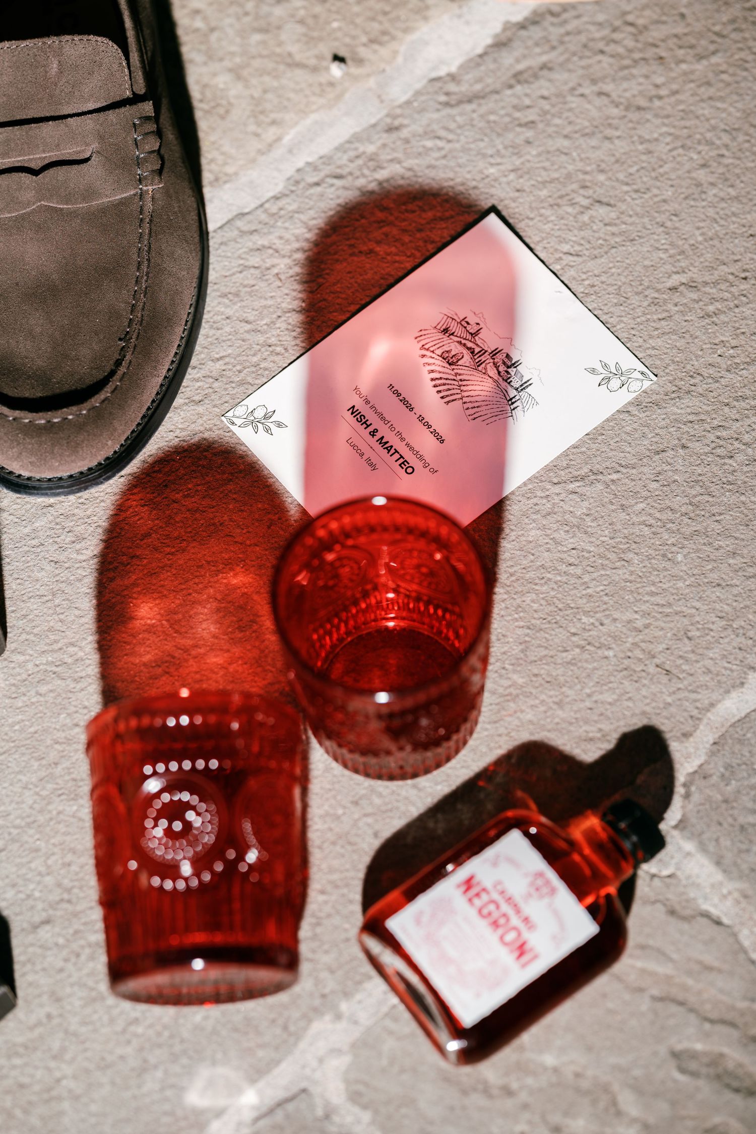 Red glass tumblers and a small Negroni bottle beside a wedding invitation and brown suede loafer on stone, casting red shadows.