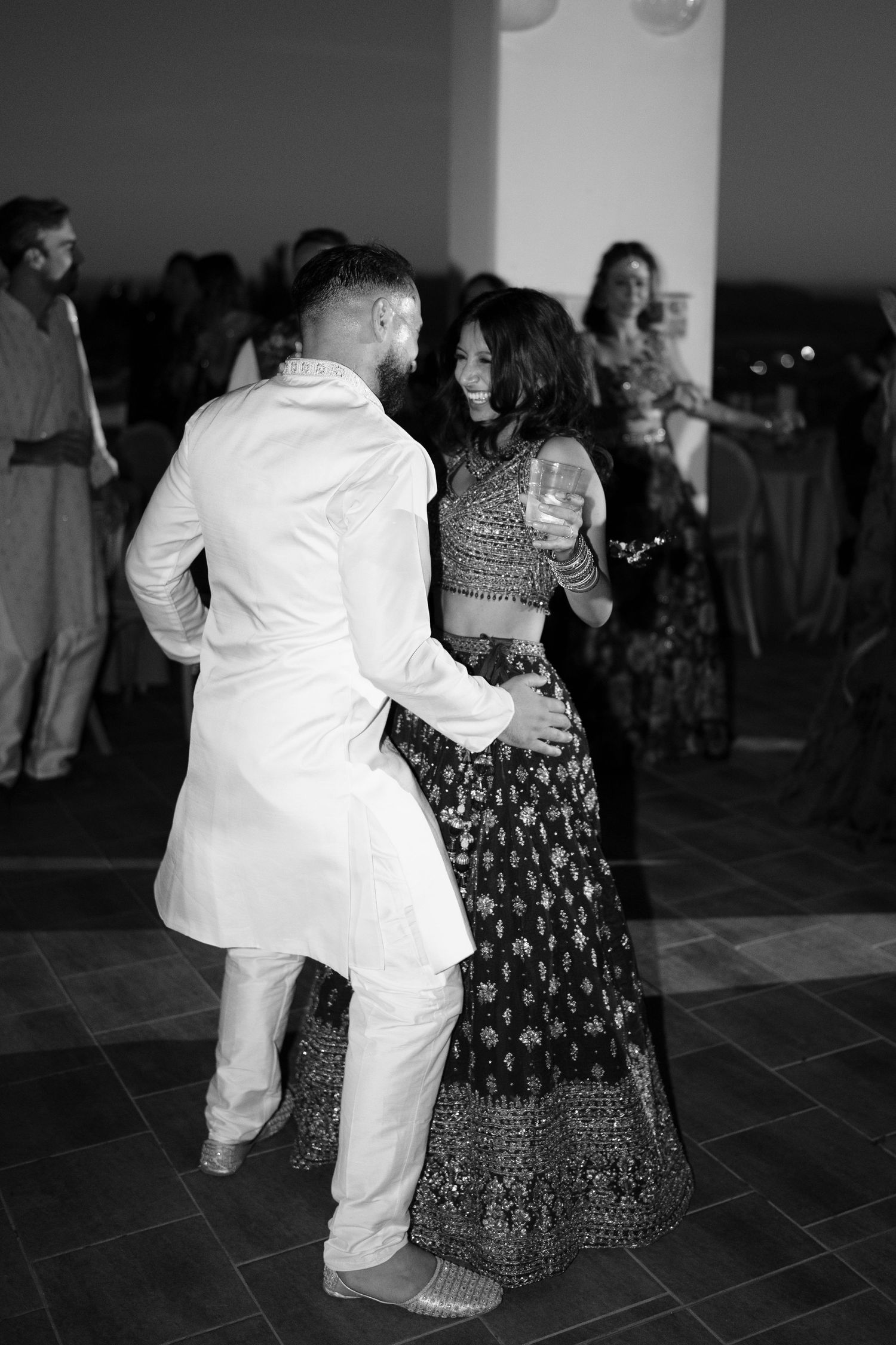 Black and white photo of a smiling couple in traditional Indian wedding outfits dancing together at an evening reception.