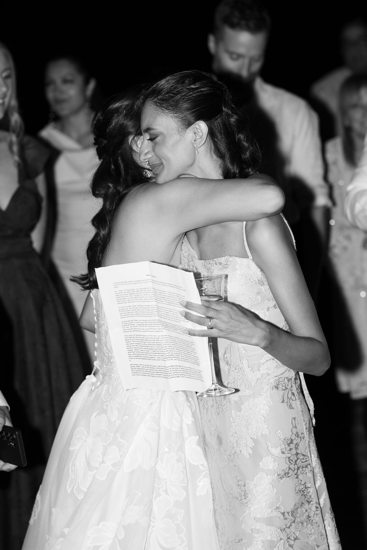 Black and white photo of a bride in a lace dress hugging a guest who holds a champagne glass and written speech at a wedding reception.
