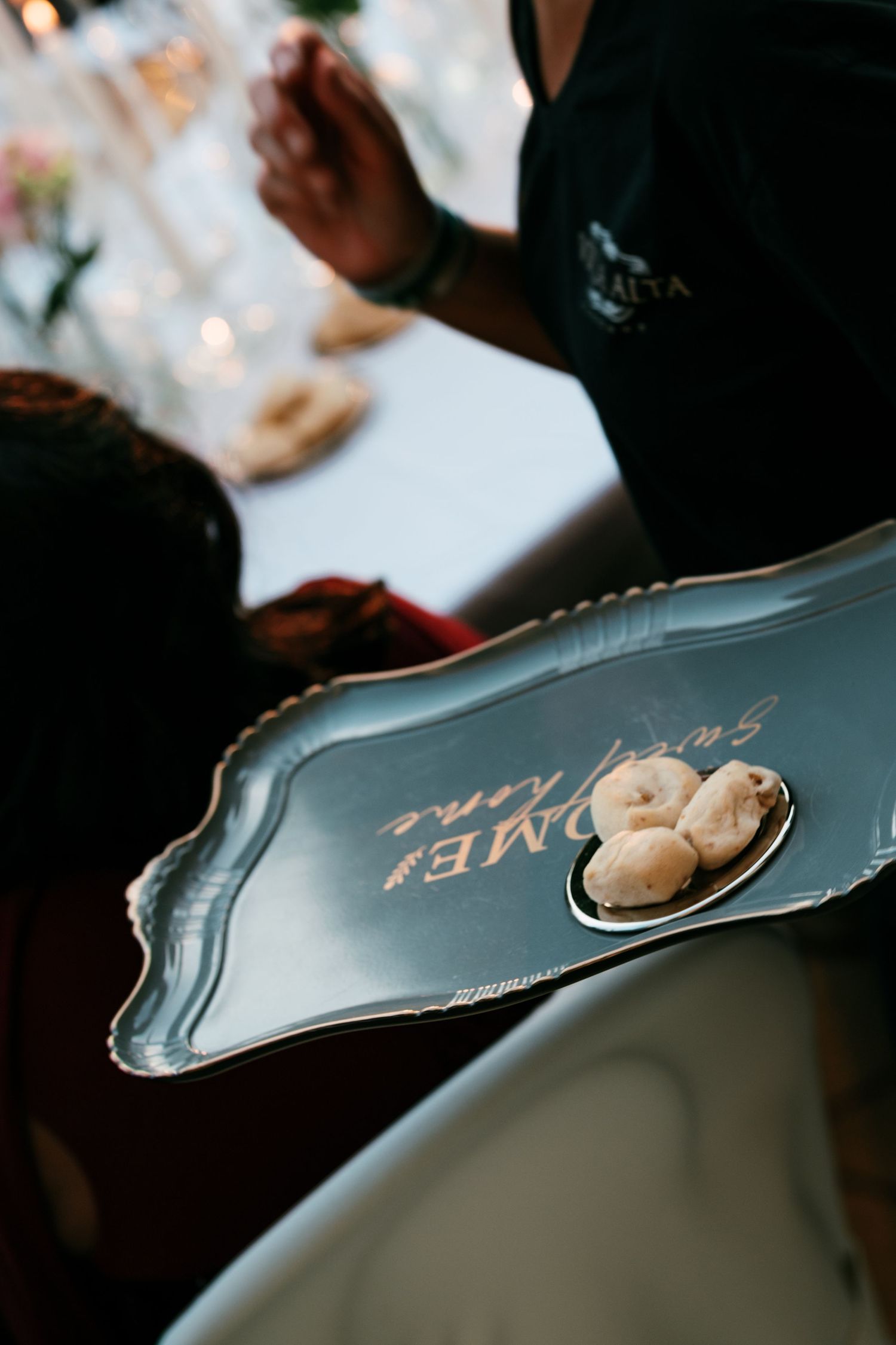 Server holding a silver tray with gold lettering that offers small pastry bites at an elegantly set candlelit table.