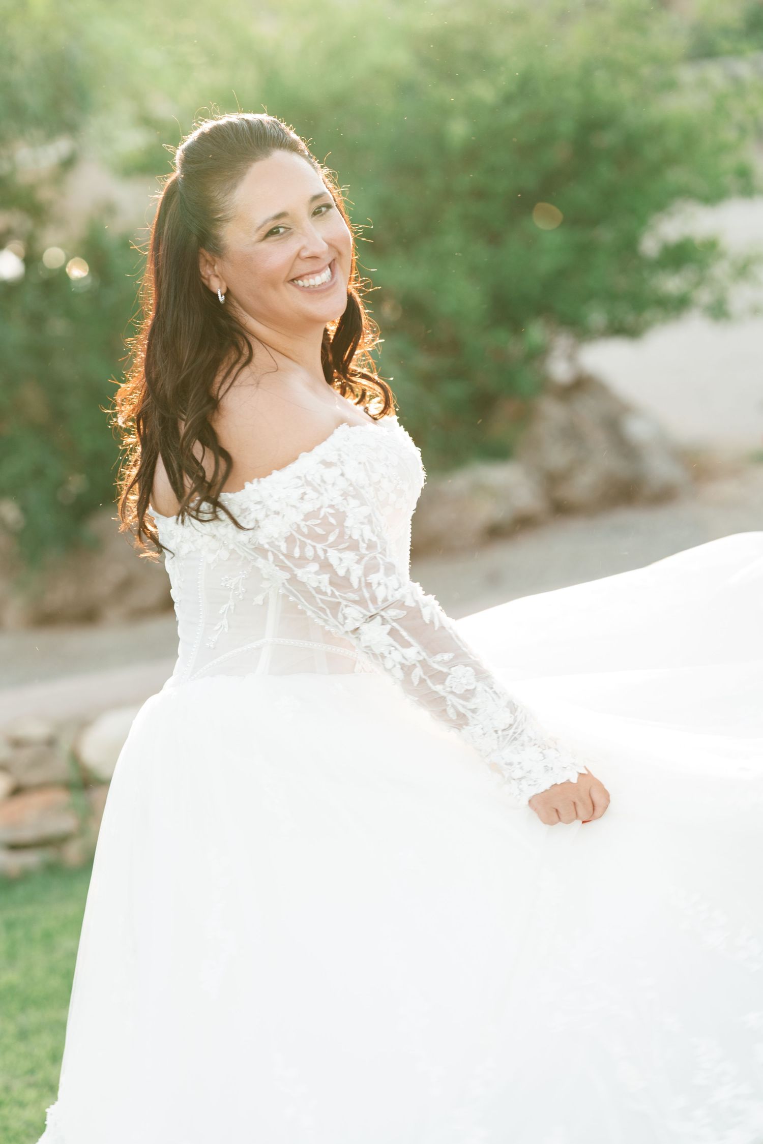 Smiling bride with long wavy dark hair twirls in a white off-the-shoulder lace wedding gown with a full tulle skirt outdoors.