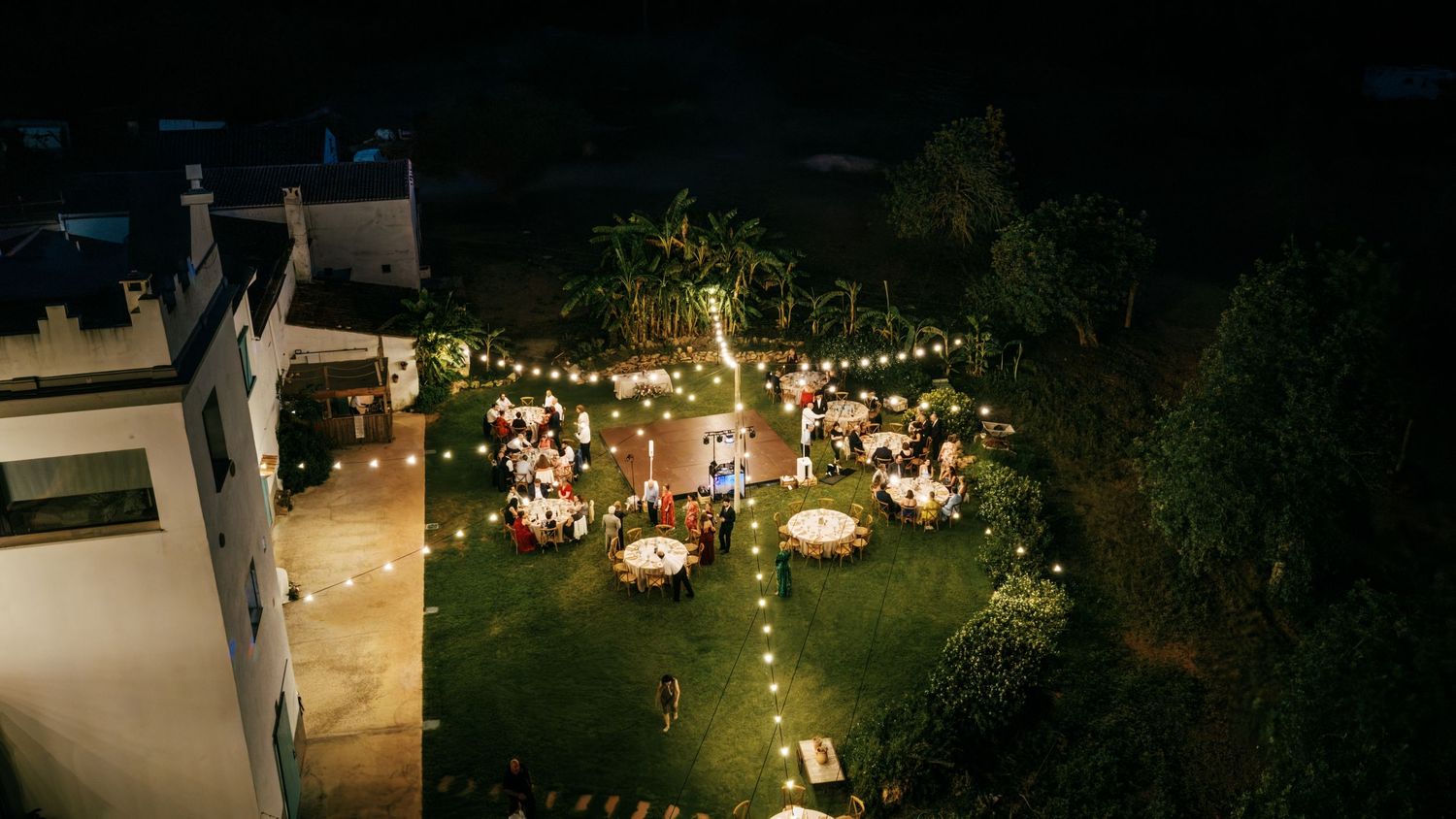Aerial view of an outdoor wedding reception on a lawn with round tables, string lights and guests beside a white building at night.