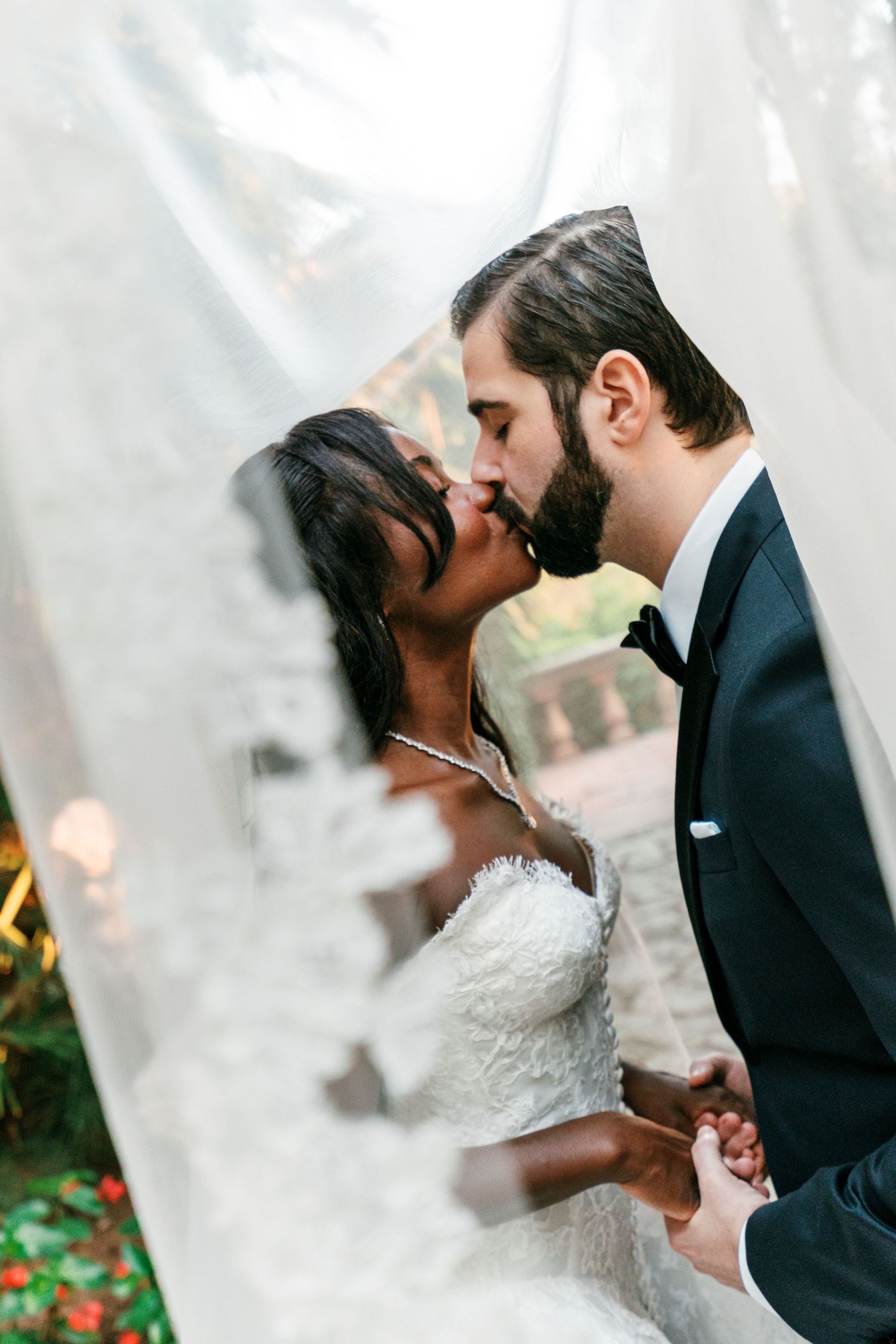 Groom in a black tuxedo and bow tie kisses his bride in a lace gown beneath a sheer veil in a garden.