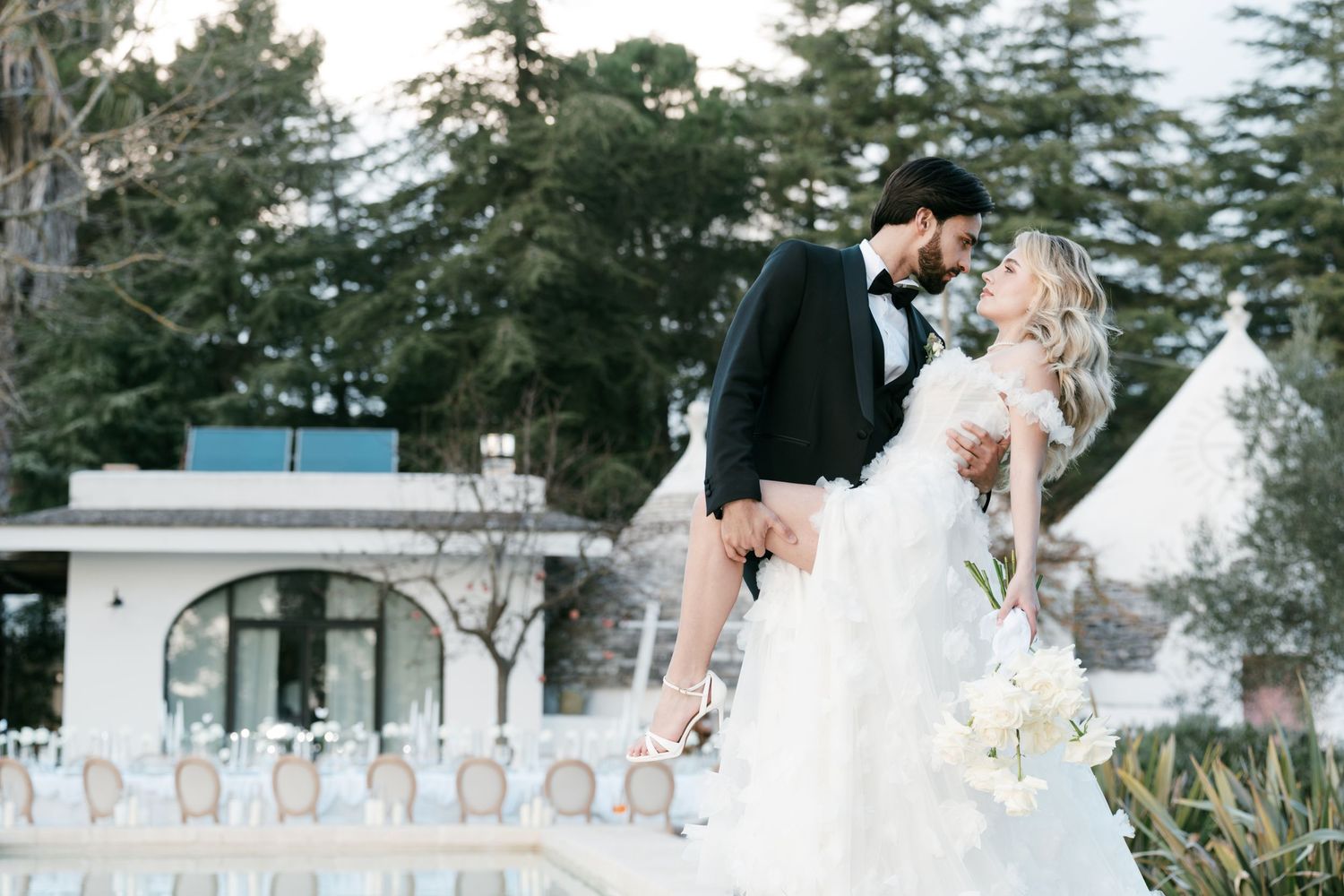 Groom in tuxedo lifts bride in voluminous white dress outdoors near elegant white venue with tall trees. Puglia wedding photographer, film