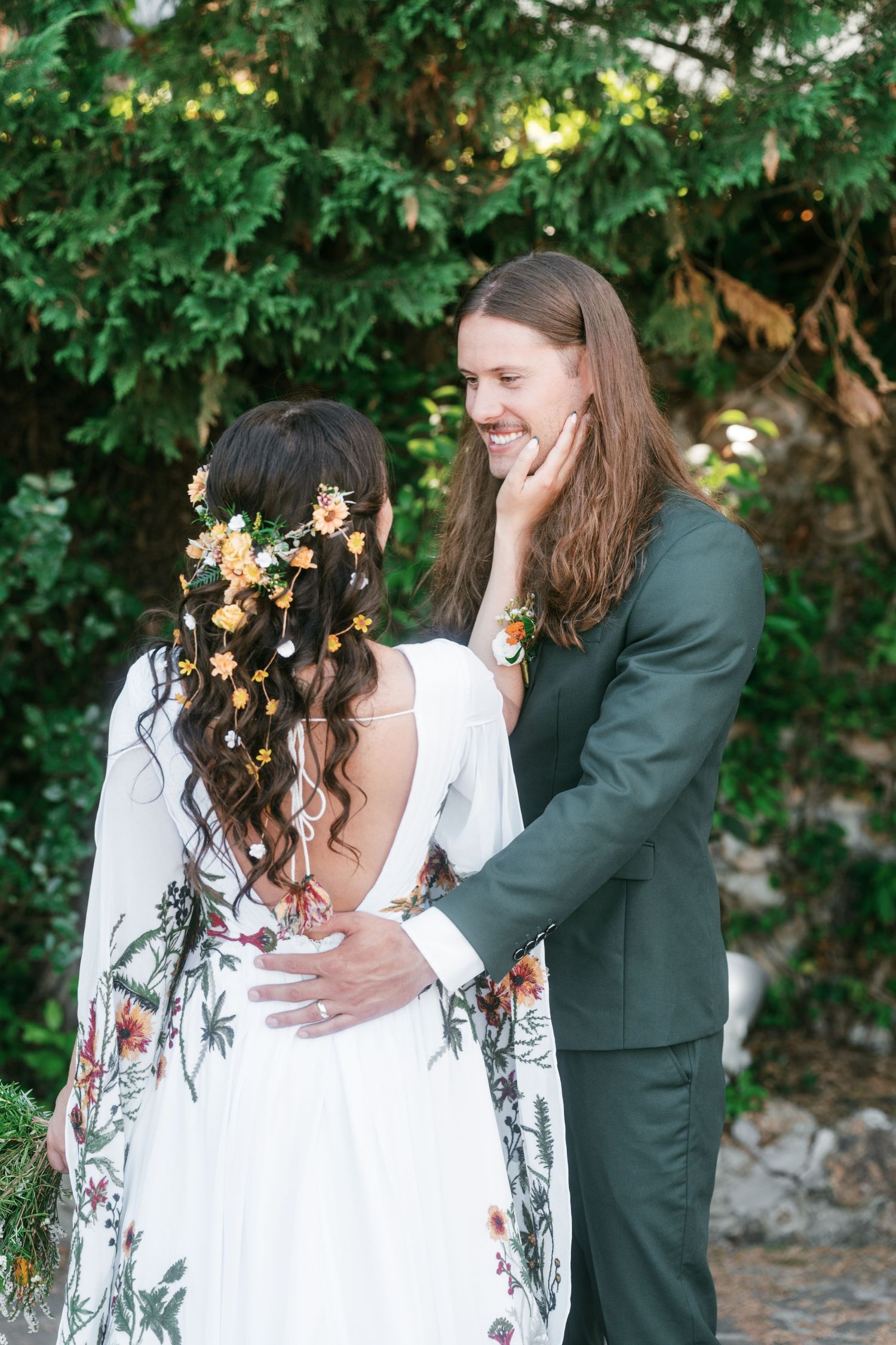 Smiling groom in green suit embraces bride in floral dress with flower crown. Tuscany wedding photographer, filmmaker