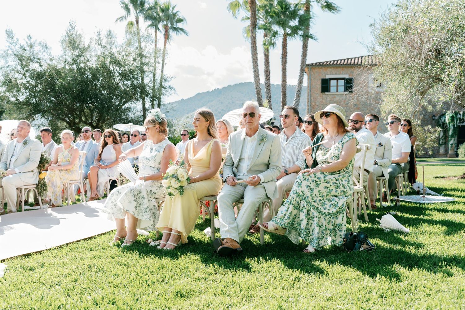 Guests seated outdoors at a sunny wedding ceremony on green lawn with Mediterranean villa and palm trees backdrop. Mallorca wedding photo