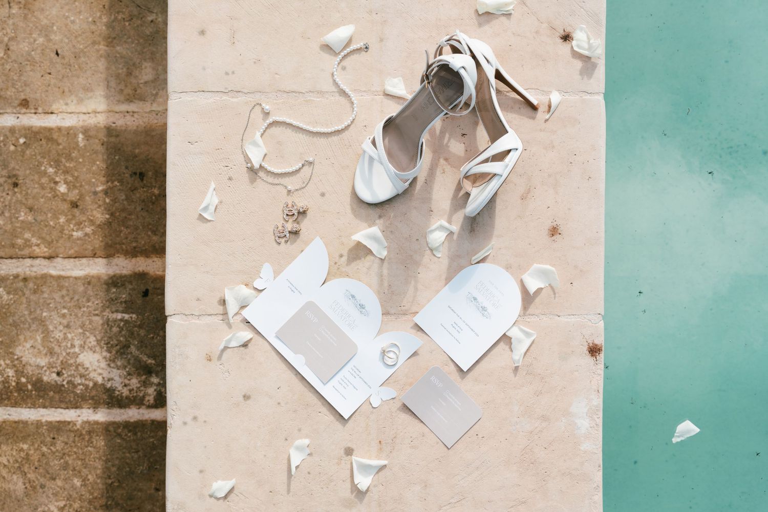 White bridal heels, wedding invitations, and accessories arranged on stone surface near pool. Puglia wedding photographer,
