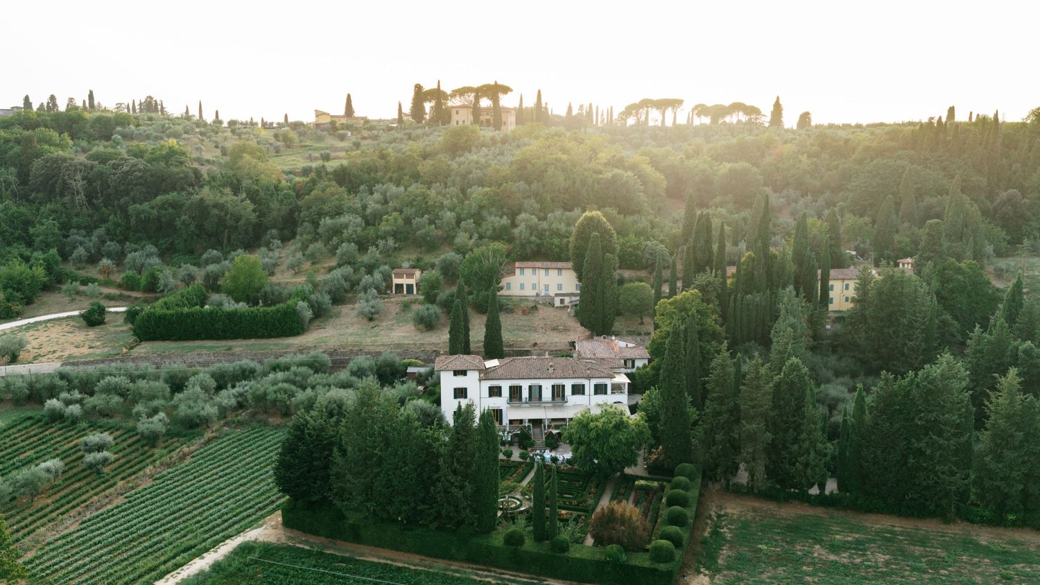 Aerial view of a Tuscan villa surrounded by cypress trees, vineyards, and rolling green hills with a distant hilltop estate at sunset.