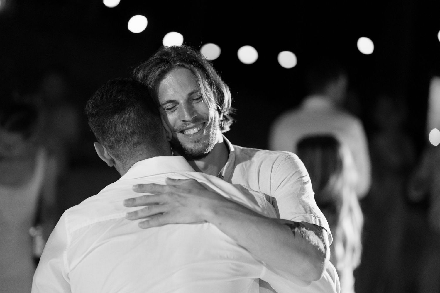 Two men sharing a warm embrace at an outdoor evening wedding celebration in black and white photography.