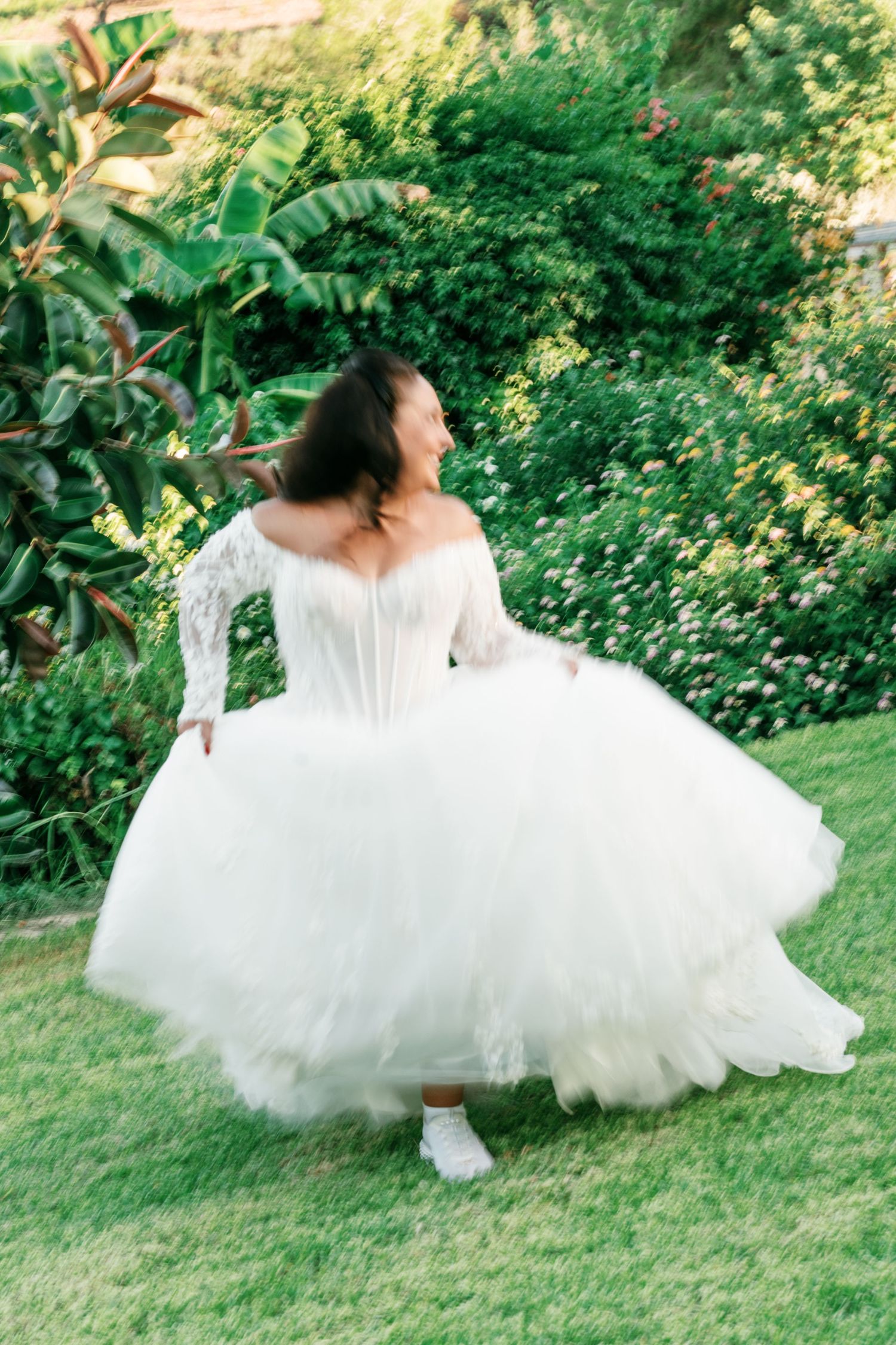Bride in a lace-sleeved corset wedding dress with a full tulle skirt twirls on a green lawn beside blooming garden flowers.
