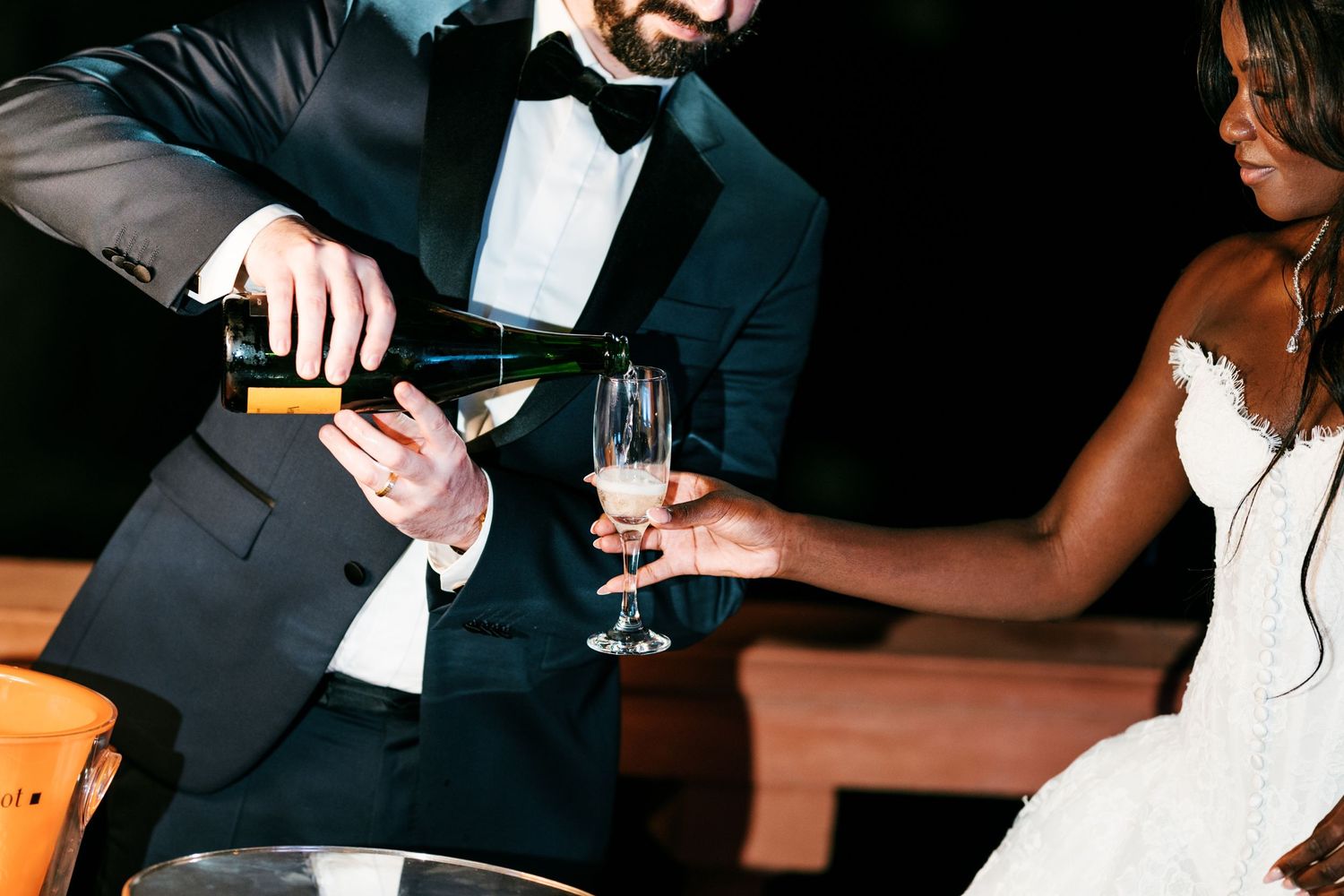 Groom in a black tuxedo pours champagne into a glass held by the bride in a white strapless wedding gown at their reception.
